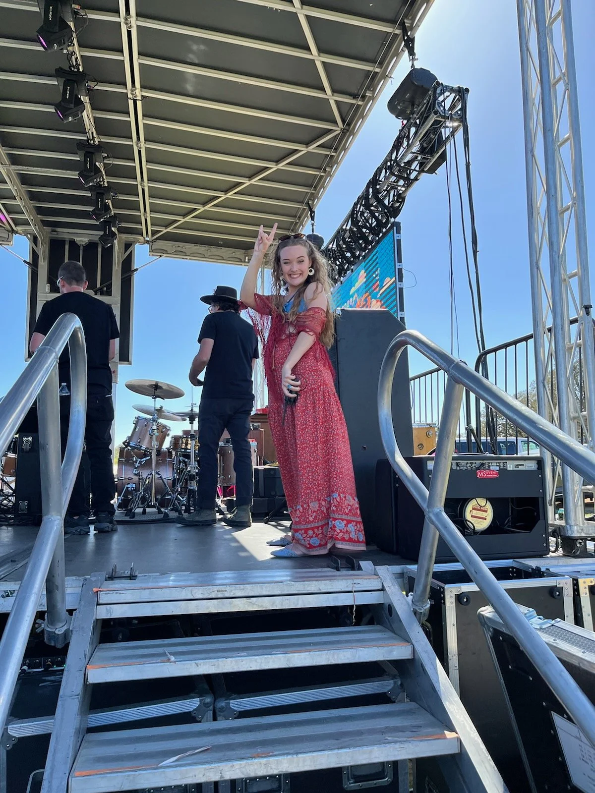 A young woman smiling and making a peace sign while standing on a stage with a microphone in her hand, wearing a red dress with floral patterns. There are musicians and stage equipment behind her, with a blue sky overhead.
