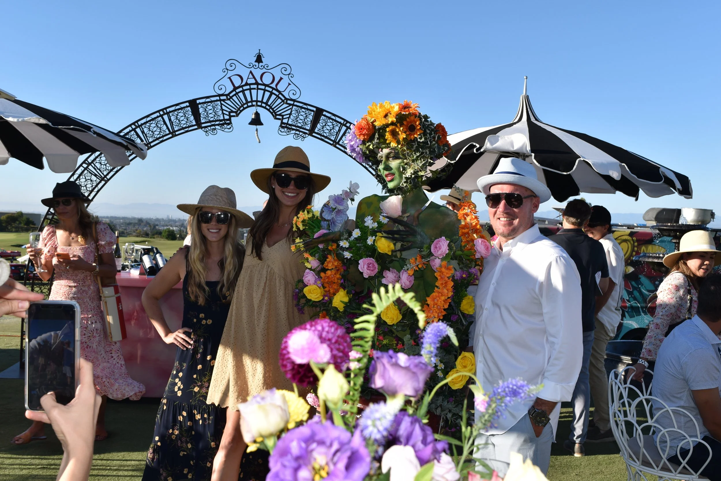People at an outdoor event, with one person dressed as a flowering woman with a flower headdress and body covered in colorful flowers, posing for photos under a decorative arch that says "DAOU," with others wearing summer dresses, hats, and sunglasses, and a scenic view in the background.