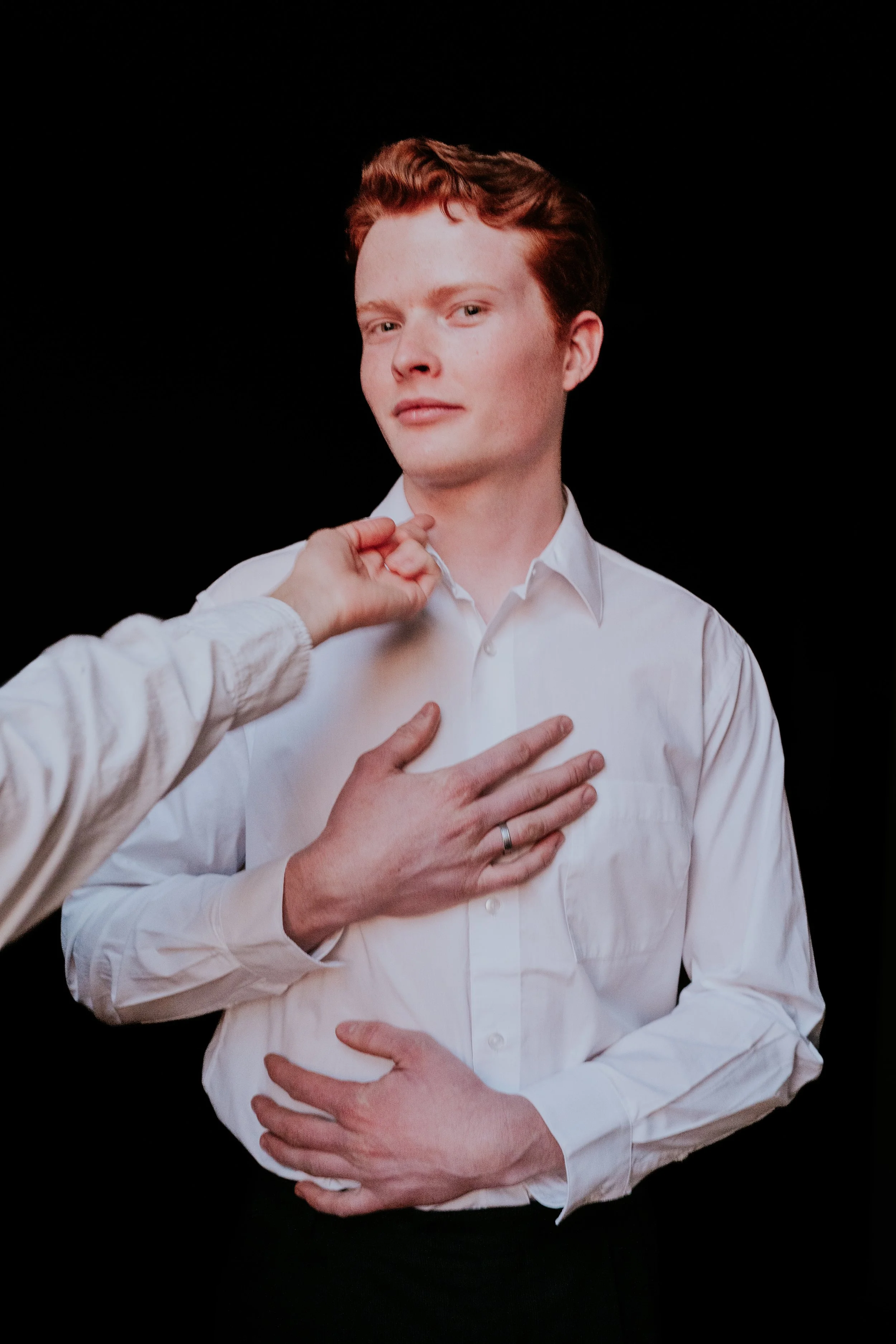 A young man with red hair wearing a white dress shirt, with two hands touching his chest and neck, against a black background.
