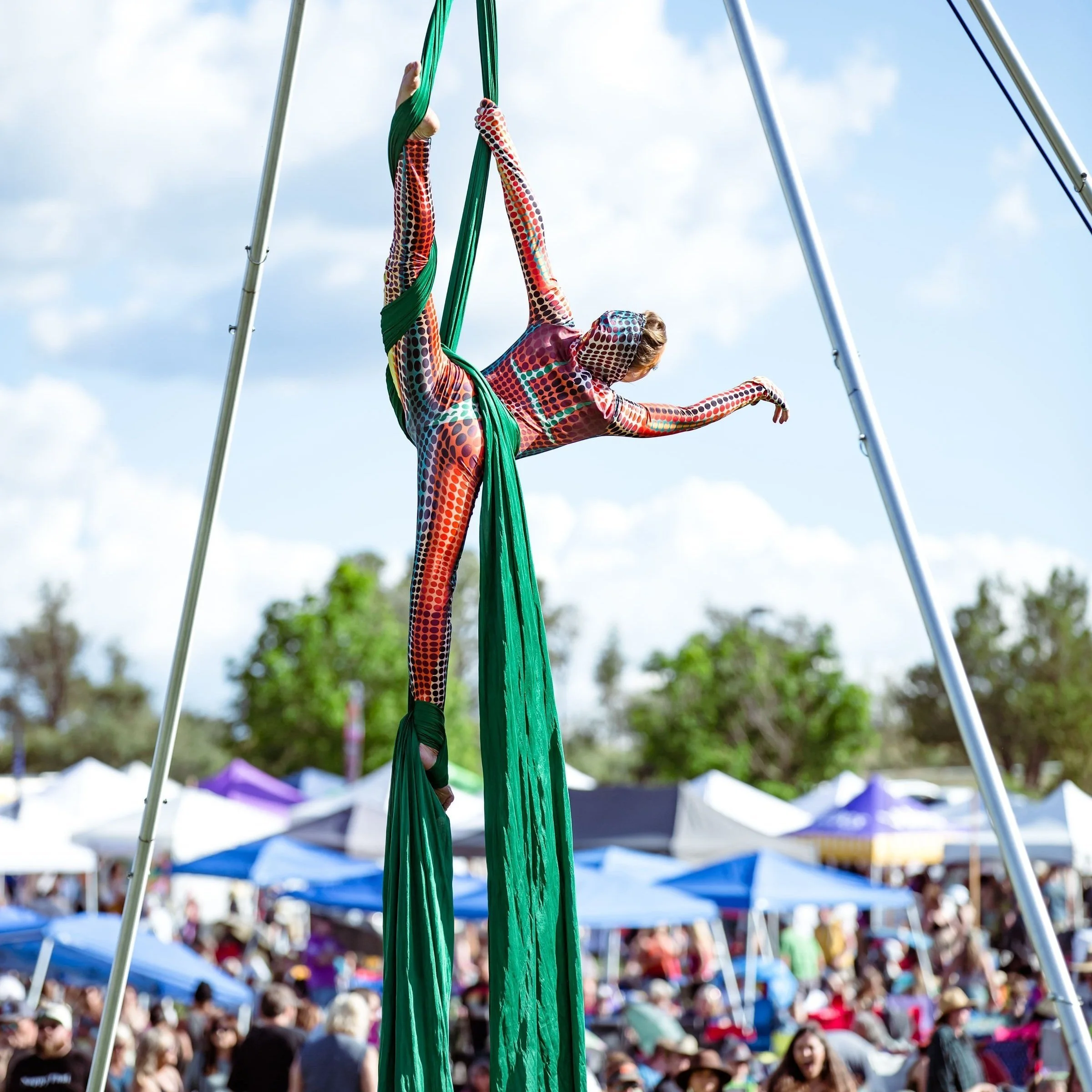 Aerialist performing on green silks outdoors during daytime with a crowd and tents in the background.
