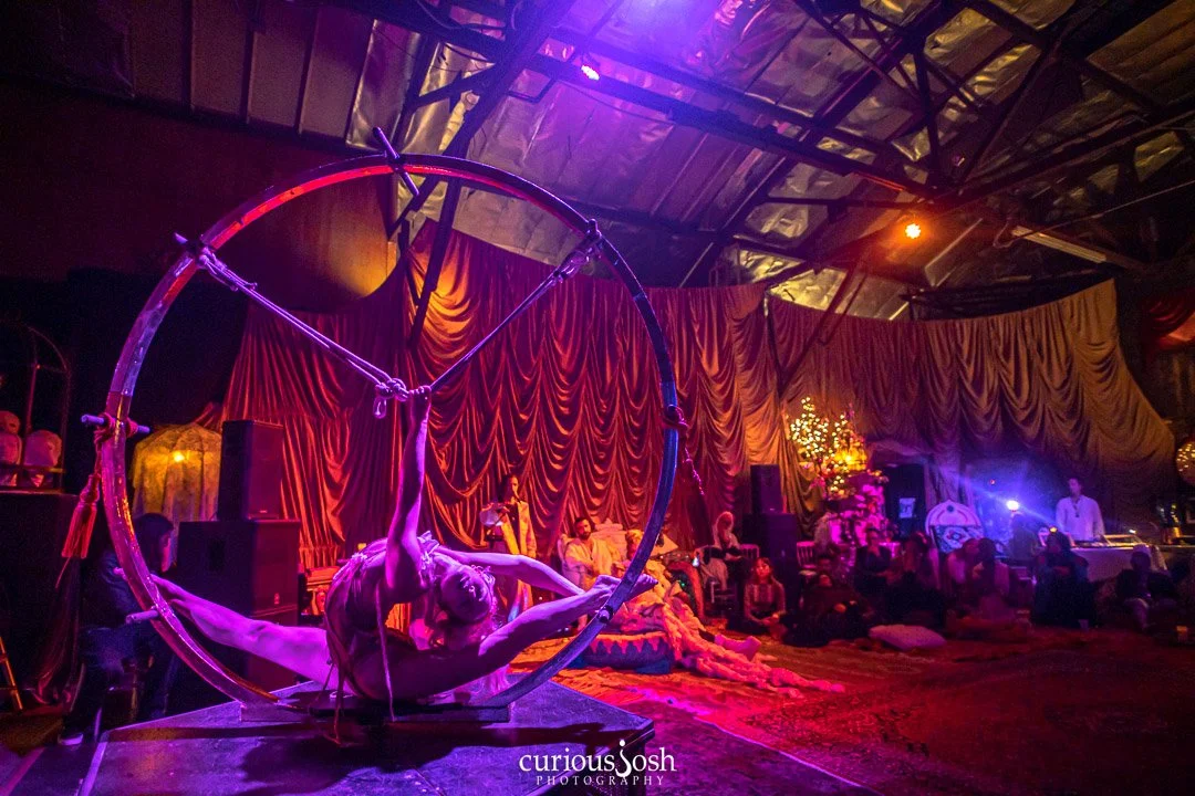 A performer on an aerial hoop in a dimly lit circus tent, with an audience seated on cushions, decorated with drapes and flowers, and colorful lighting.