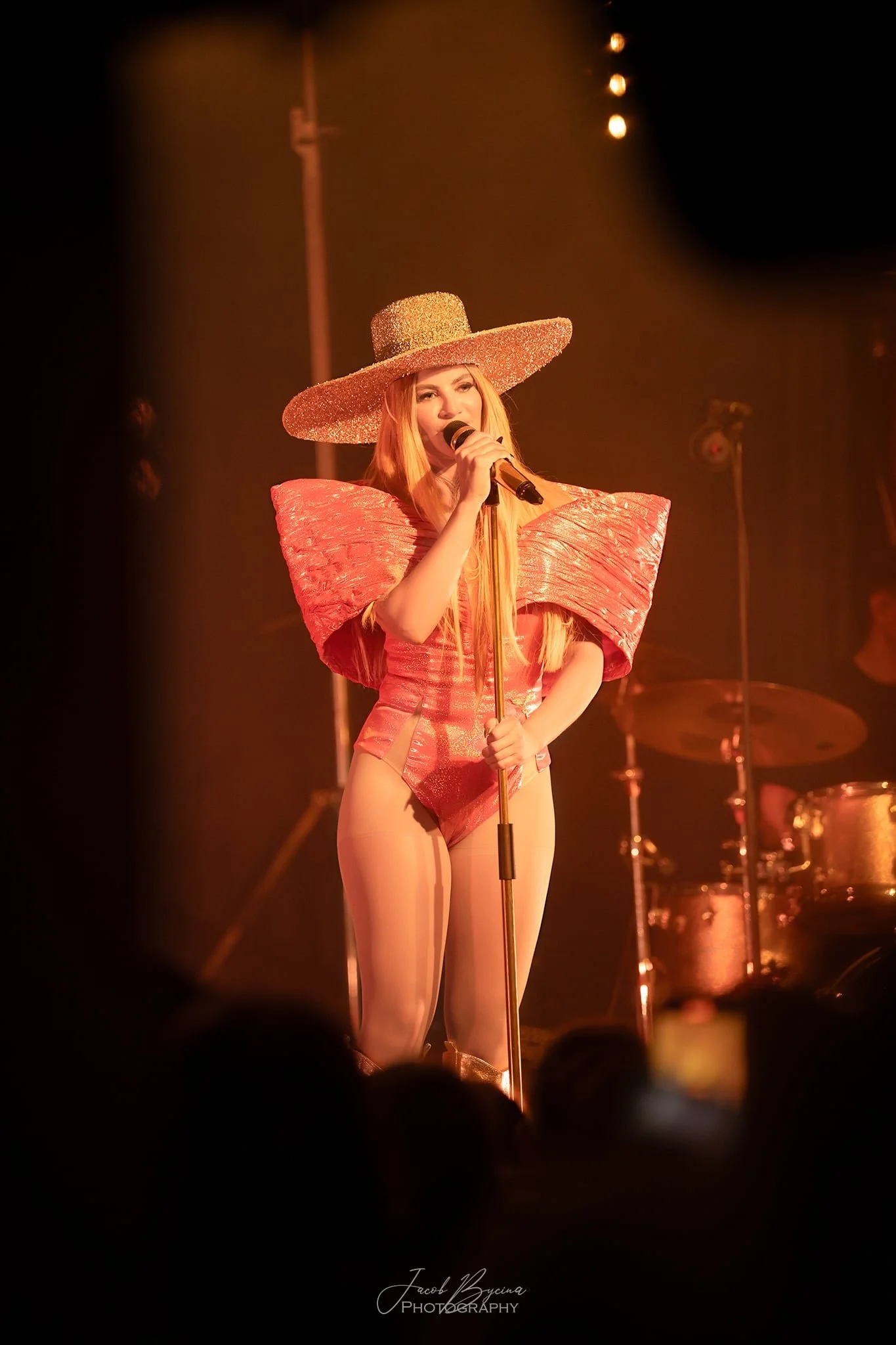A woman performing on stage wearing a large, glittery hat, a pink avant-garde outfit with structured shoulders, and sheer pink tights, holding a microphone.
