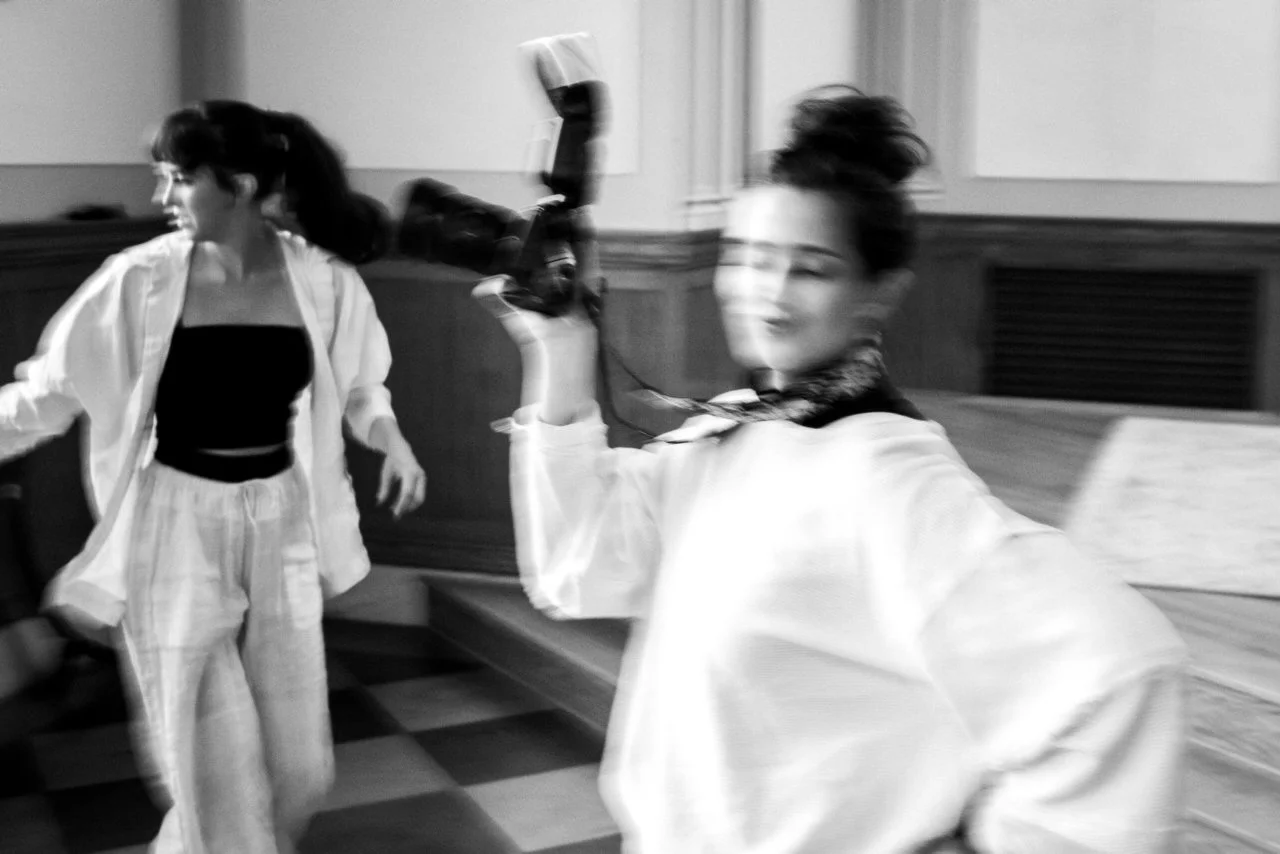 Black and white photo of two women in a room with wooden paneling. One woman is smiling and holding a camera, while the other appears to be in motion, looking away.