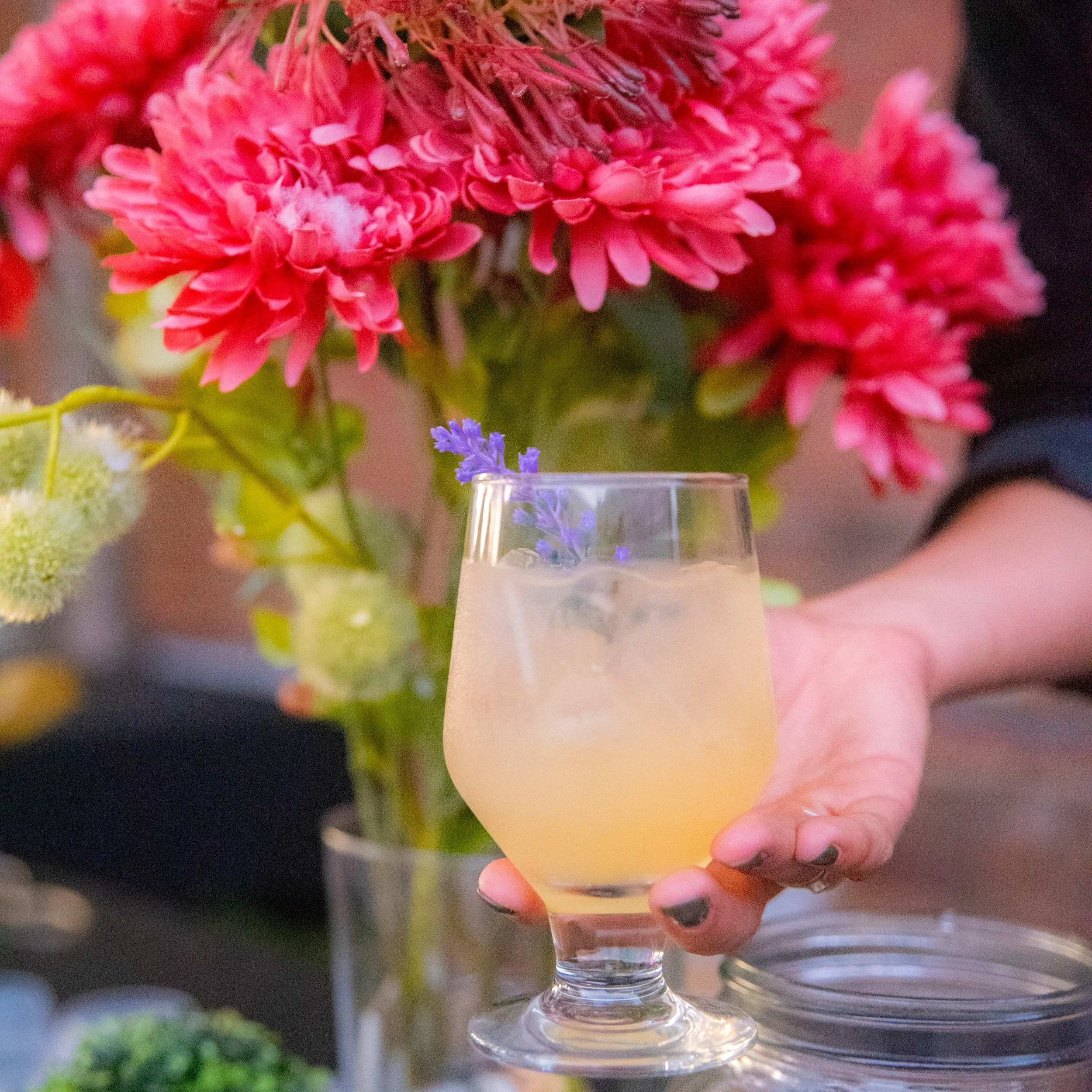 A hand holding a glass of pale yellow beverage with ice and a small purple flower garnishment, in front of a colorful bouquet of pink and purple flowers.