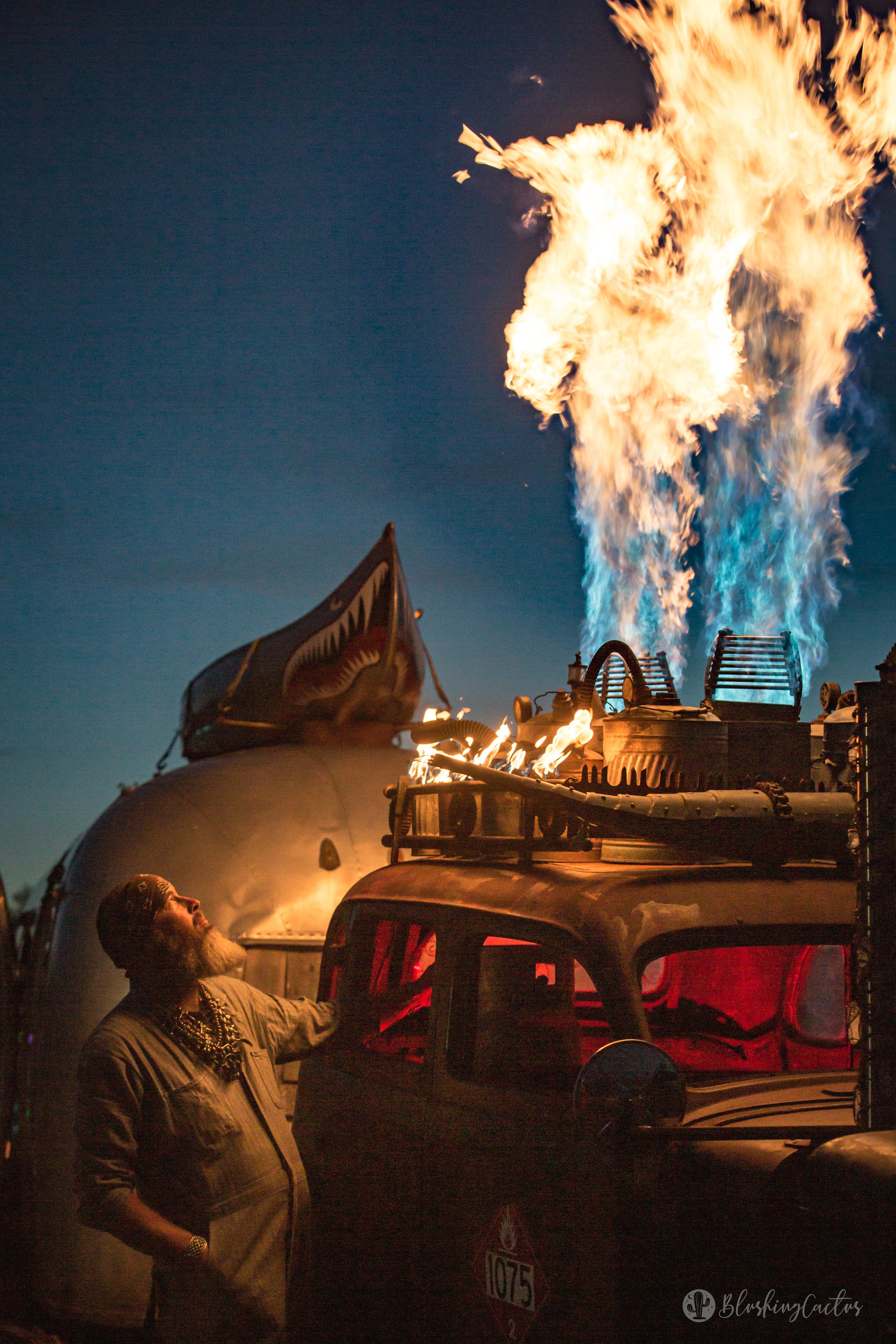 A man in a tan shirt and gold jewelry stands next to a vehicle with flames and blue fire coming from its top at night, with a flag attached to the vehicle.