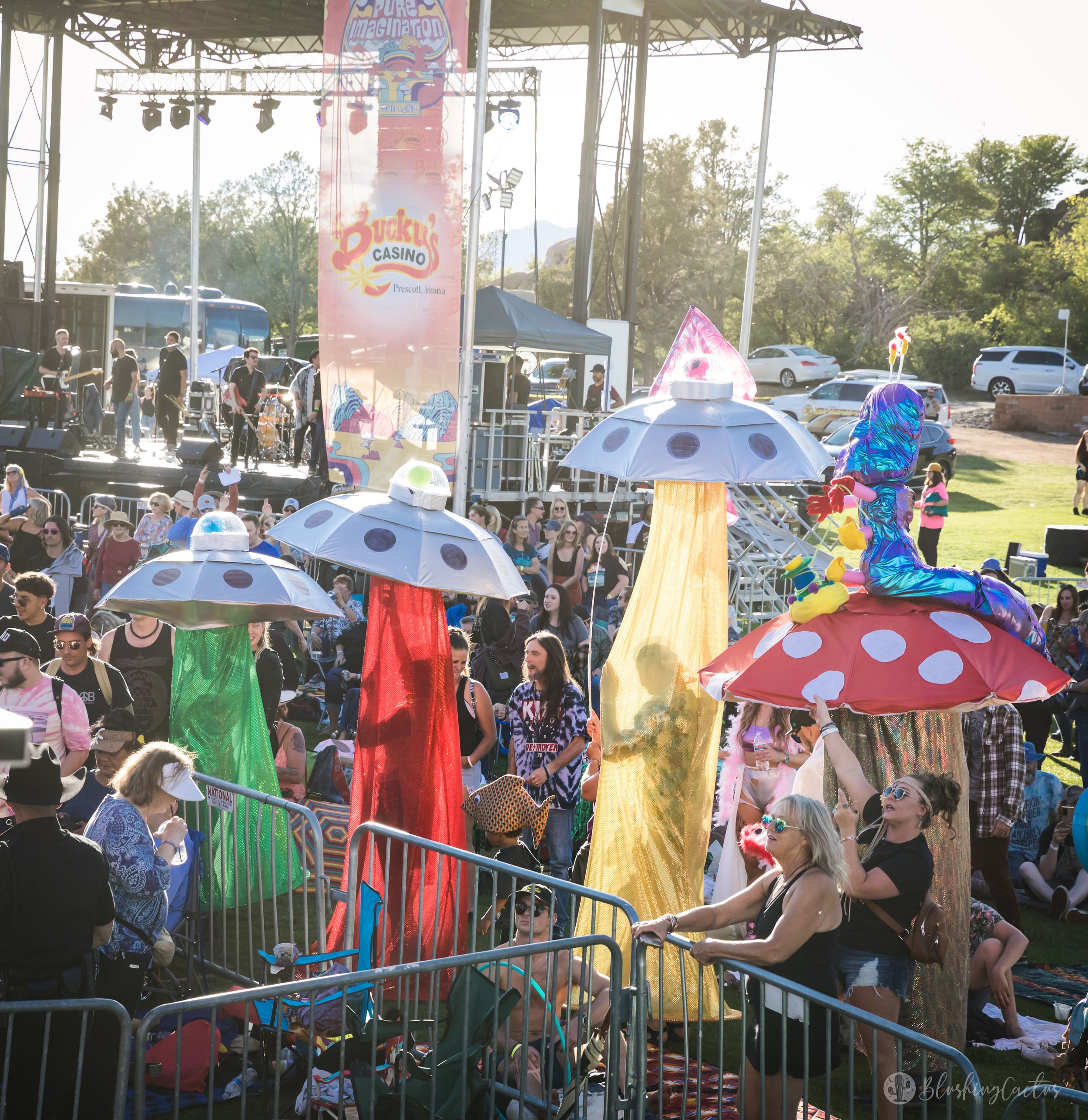 Outdoor concert with a crowd, stage in background, people sitting and standing, colorful mushroom-shaped umbrellas, and a person in a shiny, colorful costume seated on one of the umbrellas.