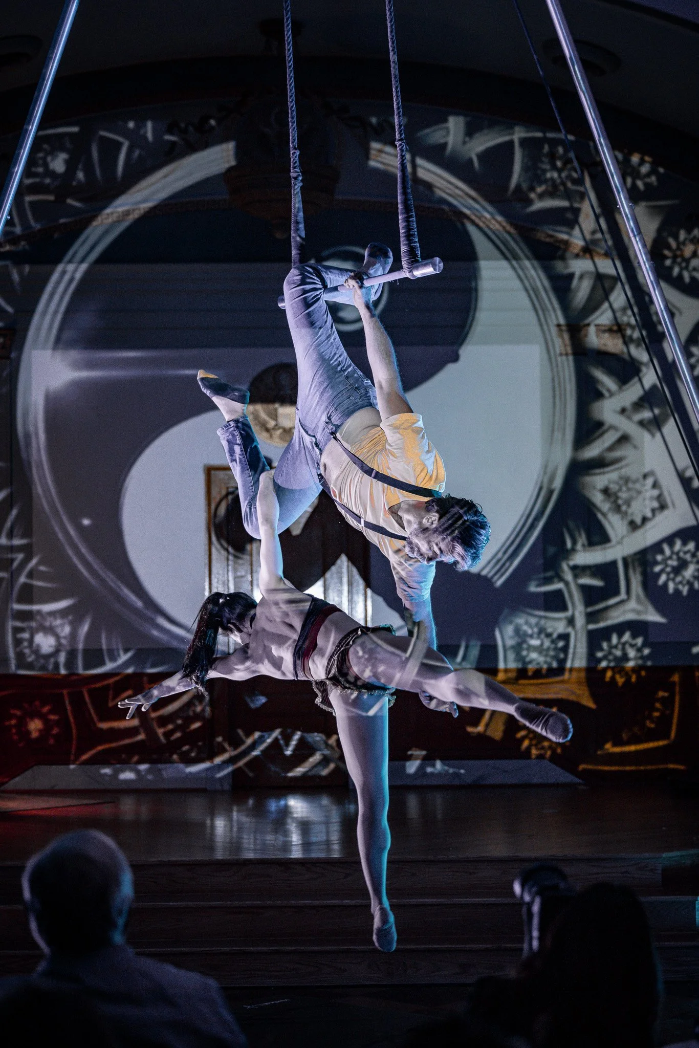 Two aerial performers demonstrate acrobatics on a swing suspended from the ceiling in a darkened theater with a projected clock background.