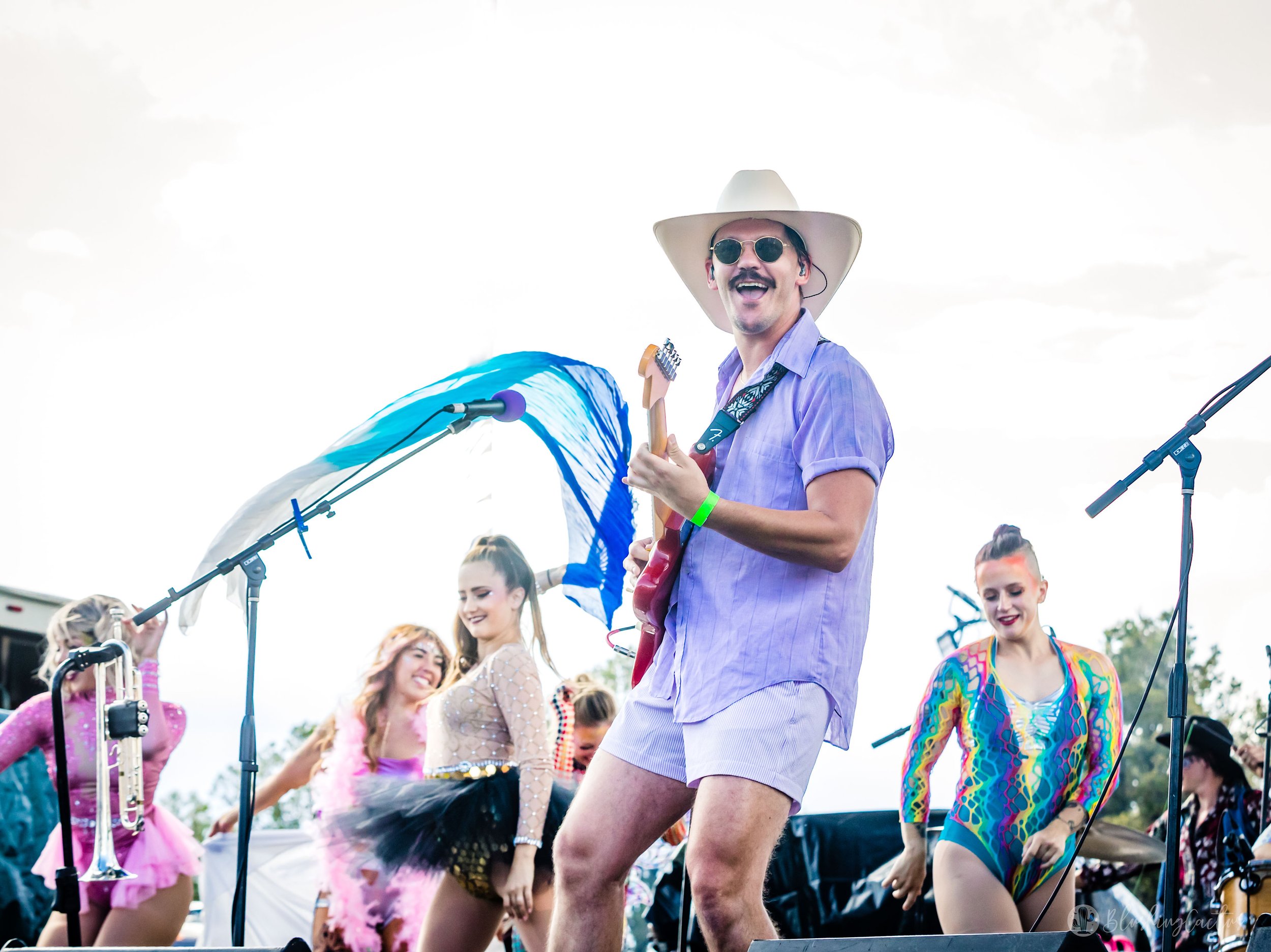 Man in a purple shirt and shorts, wearing a wide-brimmed hat and sunglasses, playing an electric guitar and singing on stage at an outdoor concert with other performers in colorful costumes.
