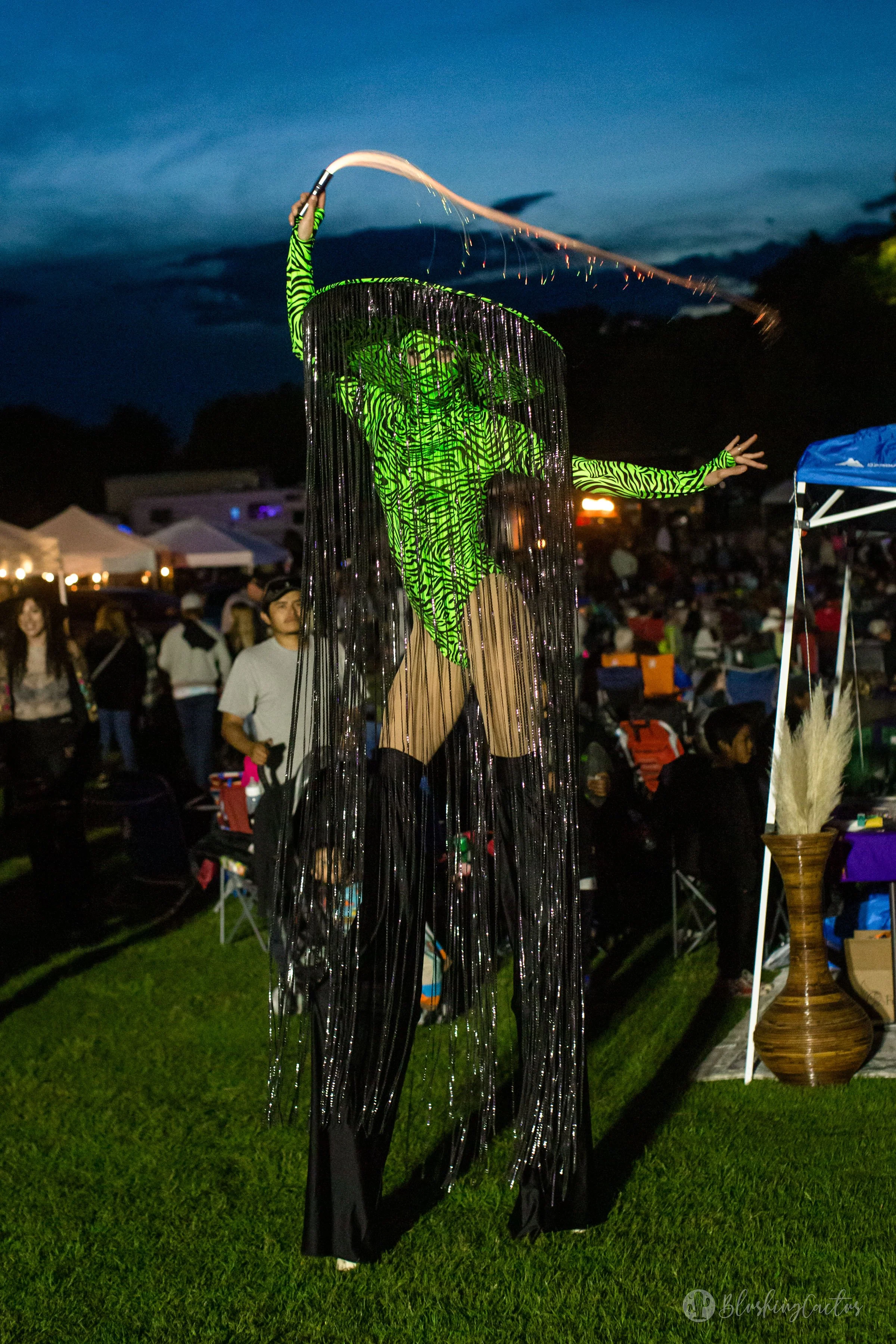 A performer on stilts wearing a bright green zebra-patterned costume and a matching mask, holding a flaming baton, with a fringe cape, performing at an outdoor event during dusk.