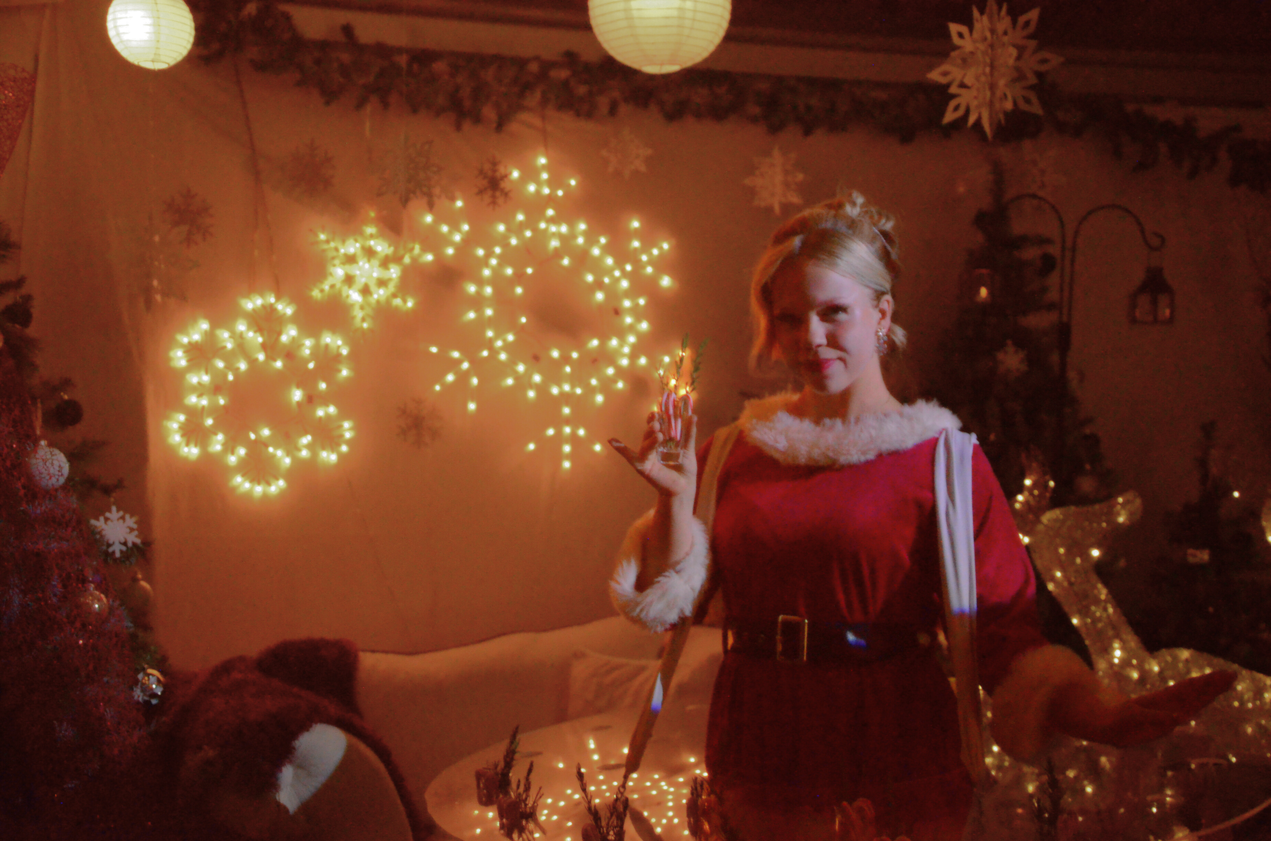 A woman dressed in a Santa outfit standing in a festive room with Christmas decorations and lights.