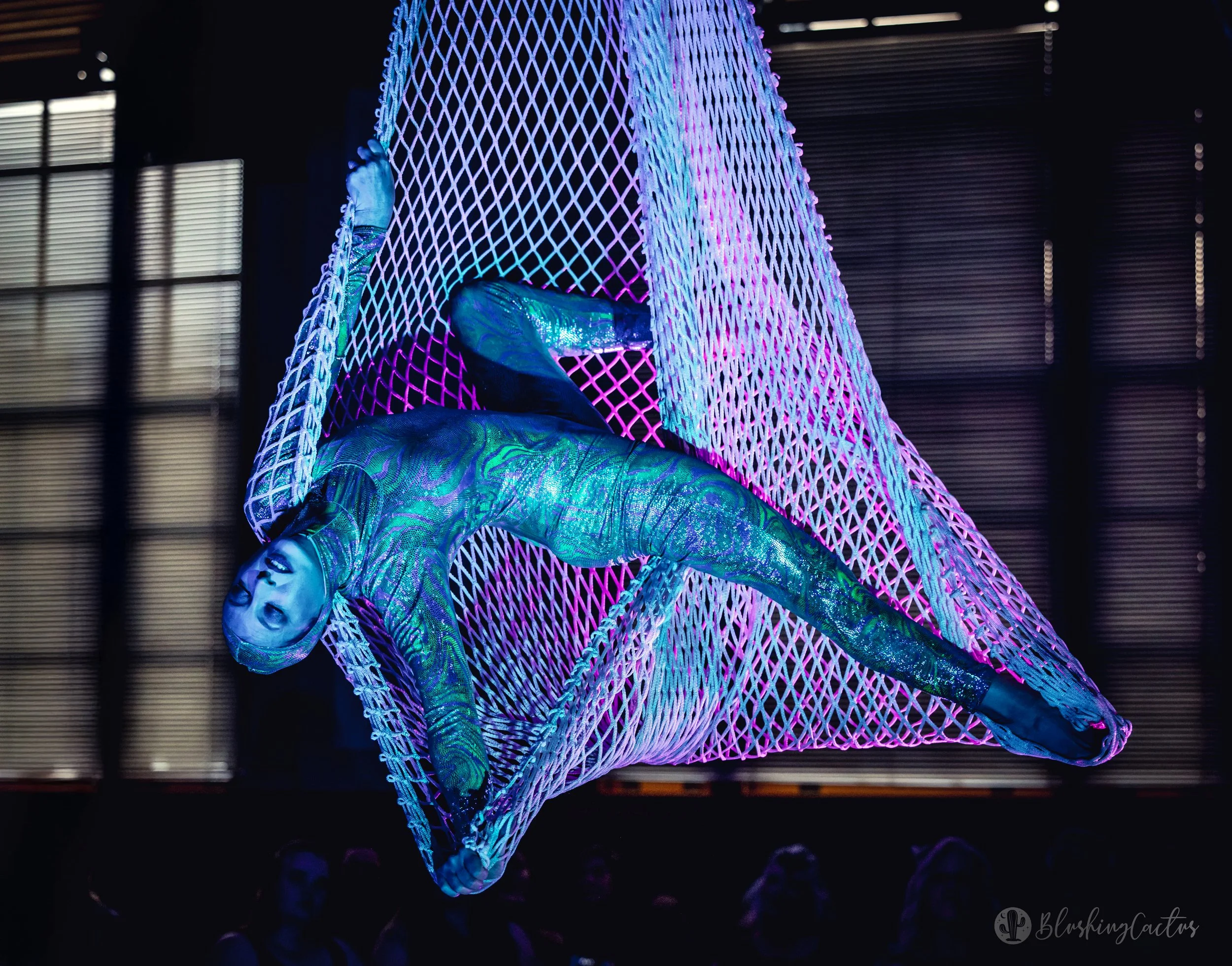 A performer in a neon-lit, rainbow-colored bodysuit with a face covering, suspended in a geometric, glowing web-like structure, against a dark background with illuminated windows.