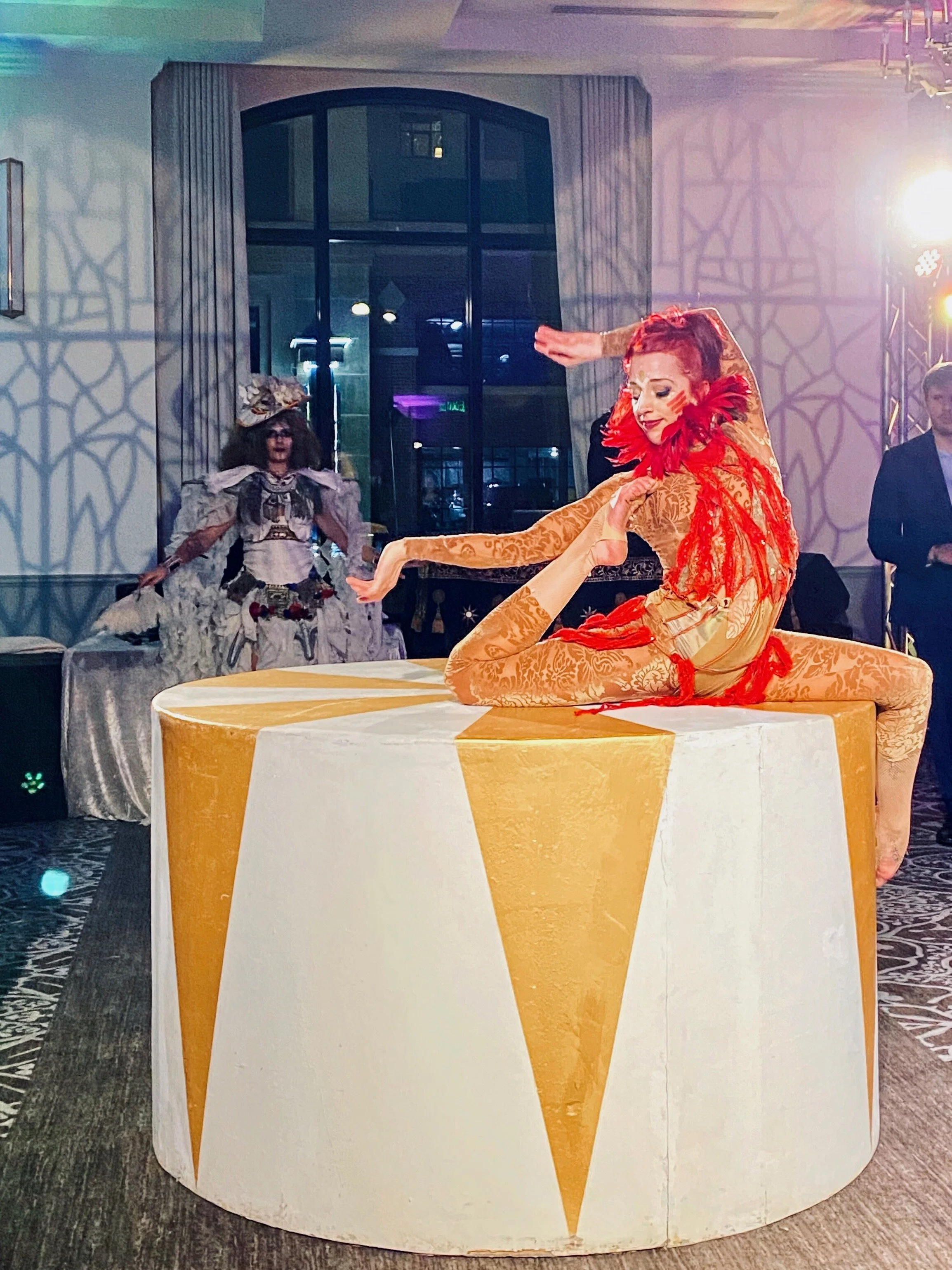 A woman with red hair in a colorful costume performing a yoga pose on a large, decorated table, with other performers and onlookers in the background at an indoor event.