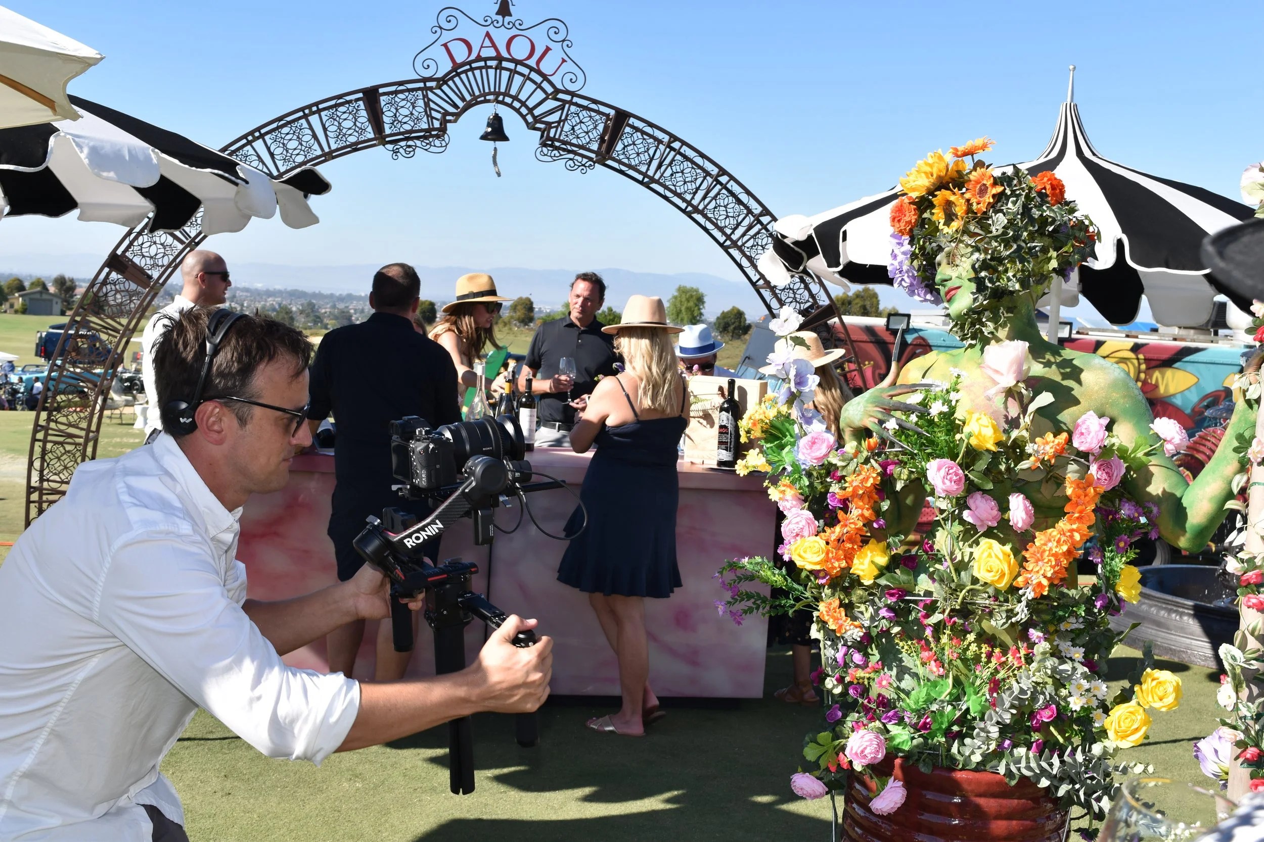 A woman with a headpiece made of colorful flowers and green body paint, flowers covering her chest and shoulders at an outdoor event. In the background, people gather around a bar under a decorative arch, with a man recording with a camera on a stabi