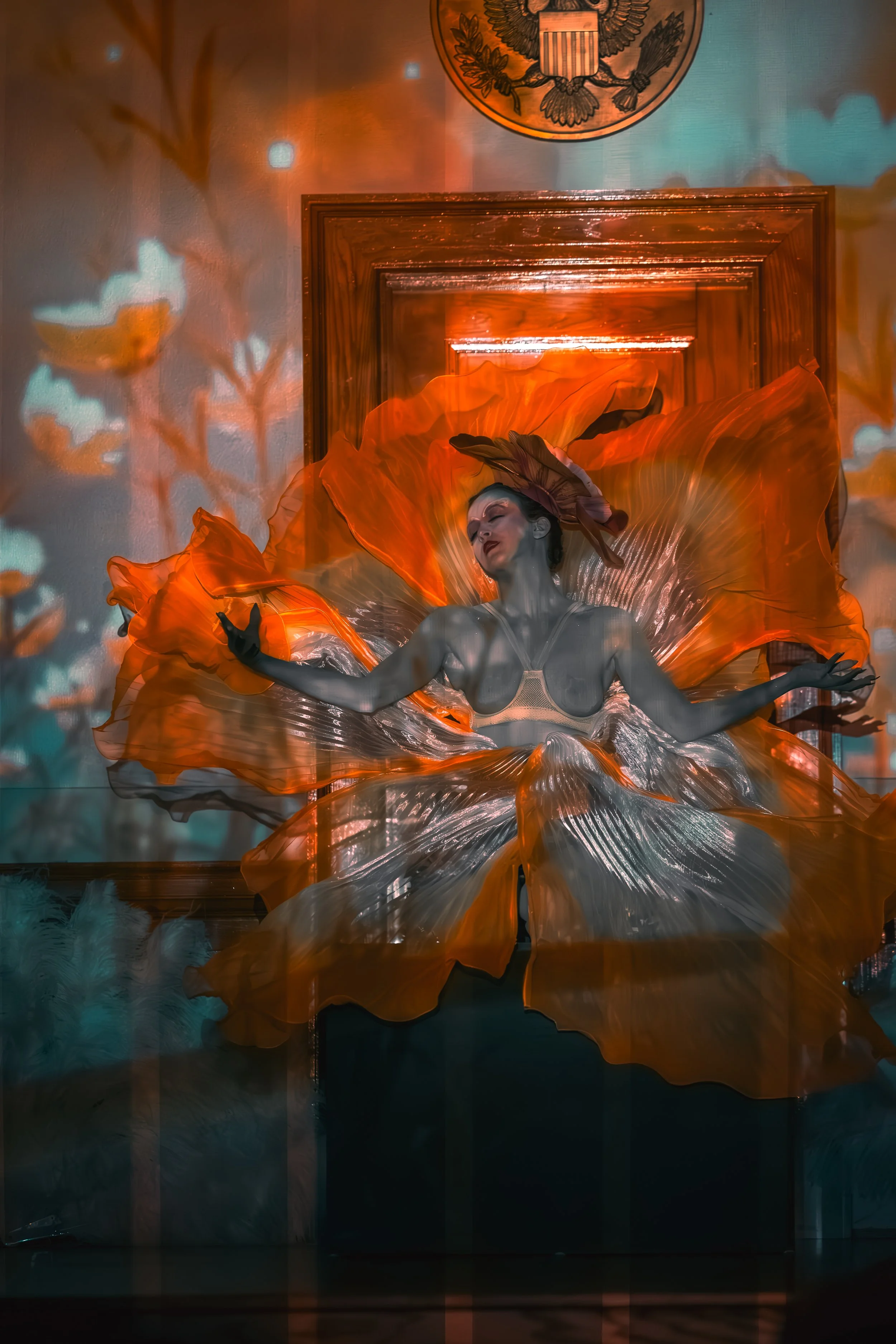 A woman in an elaborate orange and silver dress with a feathered headpiece, performing in front of a wooden door and blue wall with a crest or emblem, under colorful lighting and shadows.