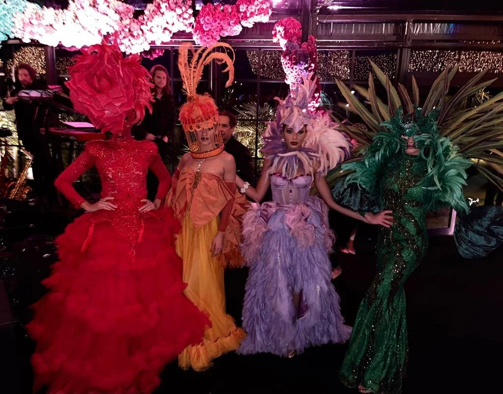 Four individuals in elaborate, colorful costumes with feather and floral embellishments pose in a decorated indoor setting.