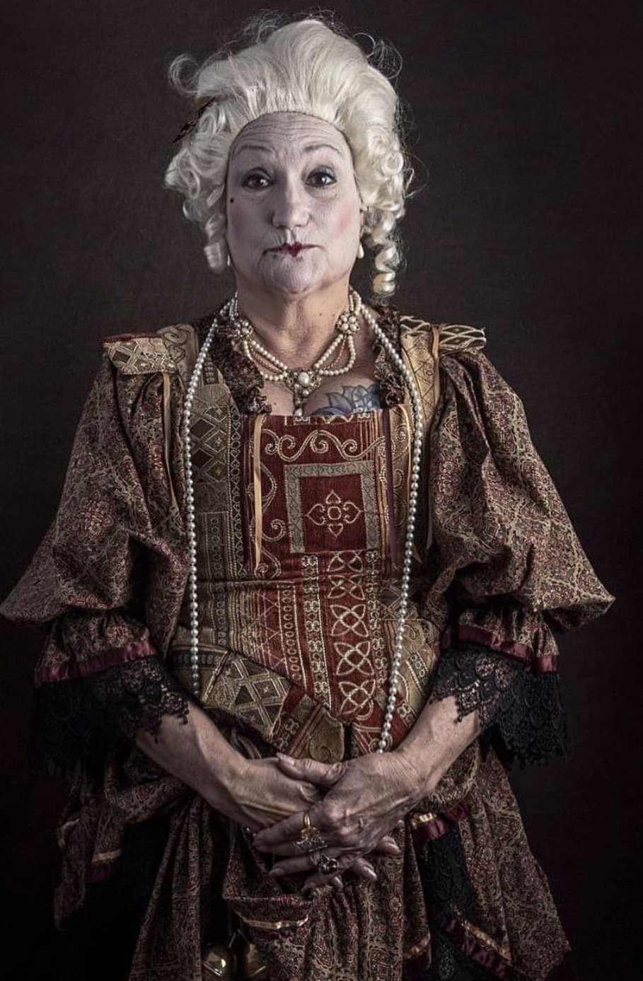 An elderly woman dressed as a regal queen, wearing a richly patterned, dark-colored gown with lace cuffs, layered pearl necklaces, and jewelry. She has styled platinum blonde hair in an elaborate fashion and is standing against a plain dark backgroun
