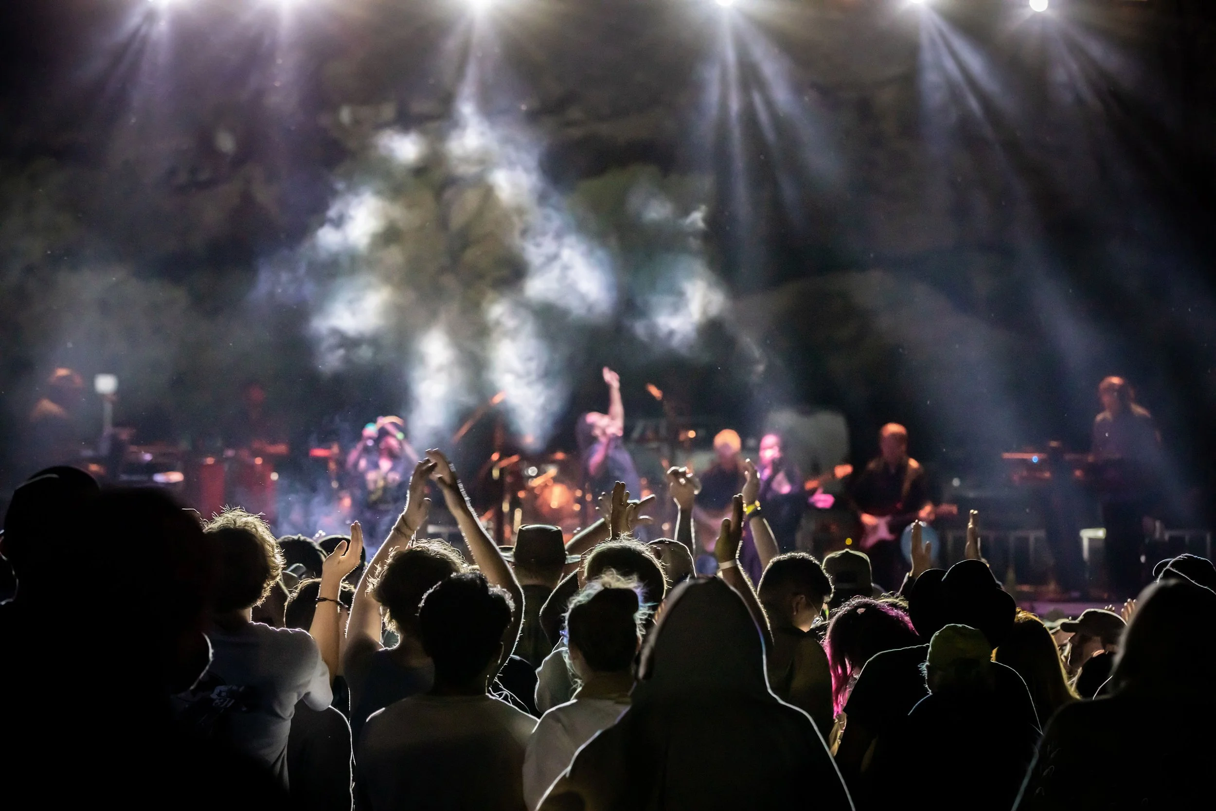 Crowd of people at a concert or music festival, facing a stage with performers and bright lights, with some raising their hands in the air, and smoke or fog creating a vibrant atmosphere.