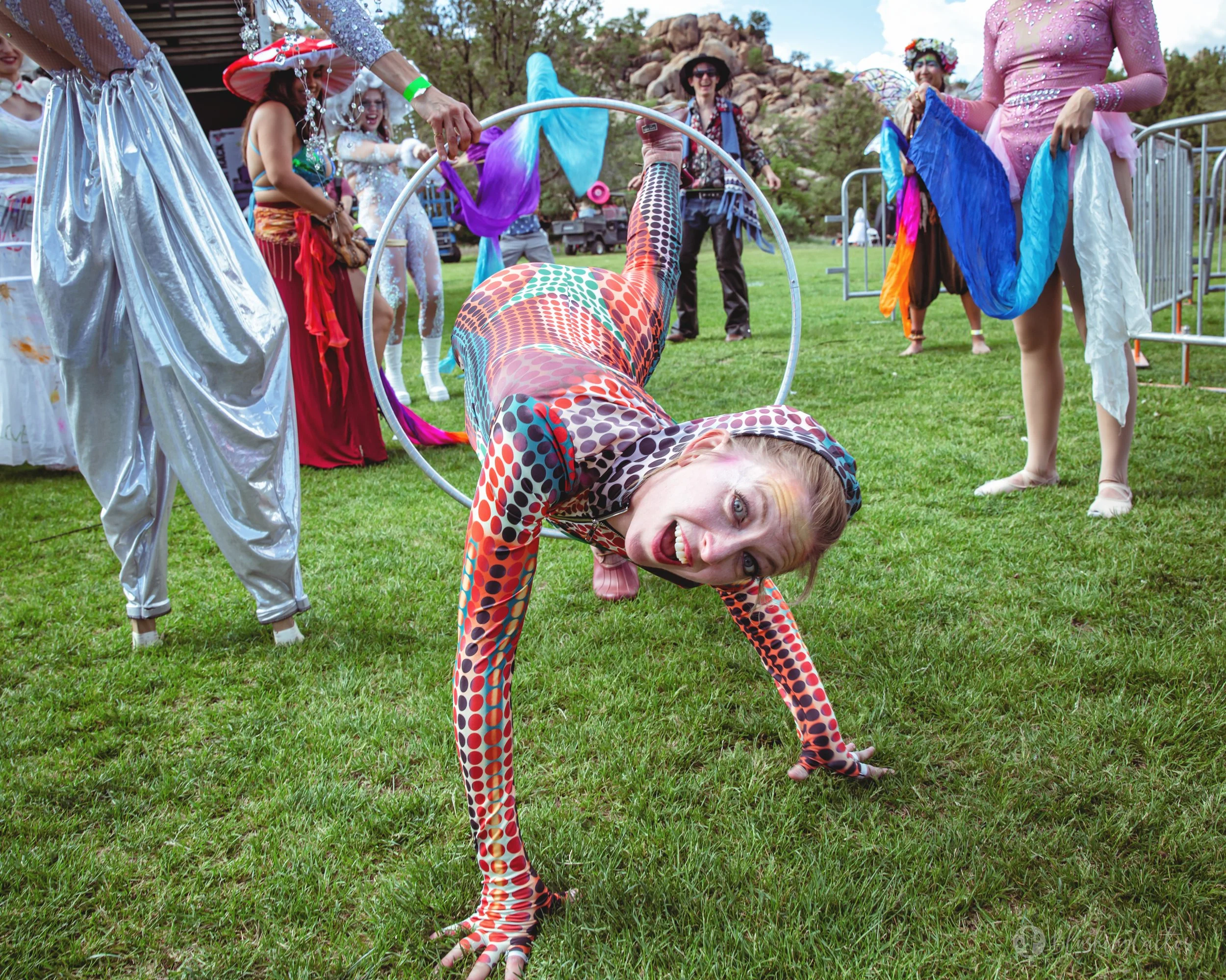 A woman dressed in a colorful, patterned bodysuit is crawling on the grass through a hoop, smiling playfully at the camera during a costume event with other women in colorful costumes in the background.
