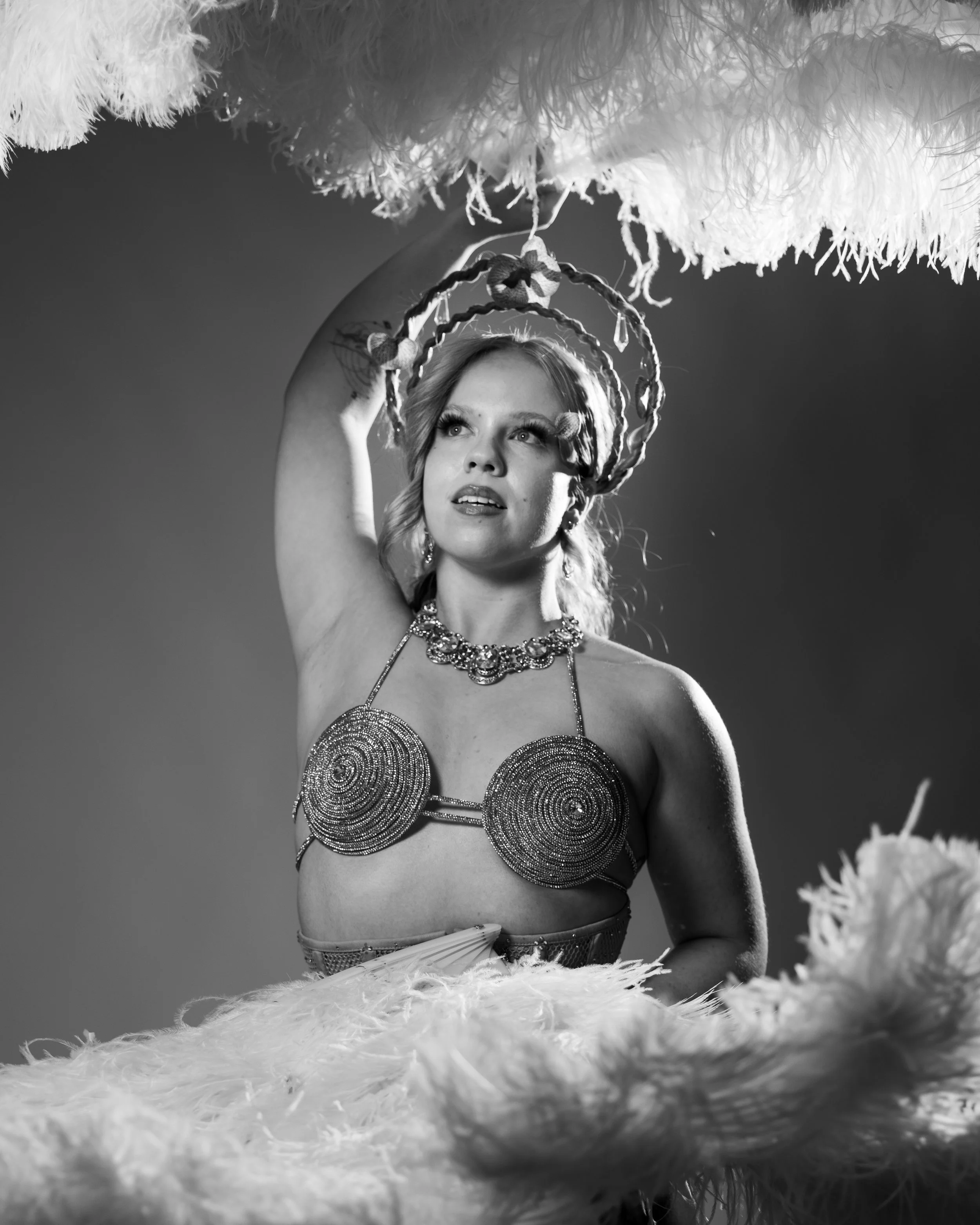 Black and white photo of a woman wearing an elaborate necklace and decorated pasties, holding a fluffy feathered object above her head.