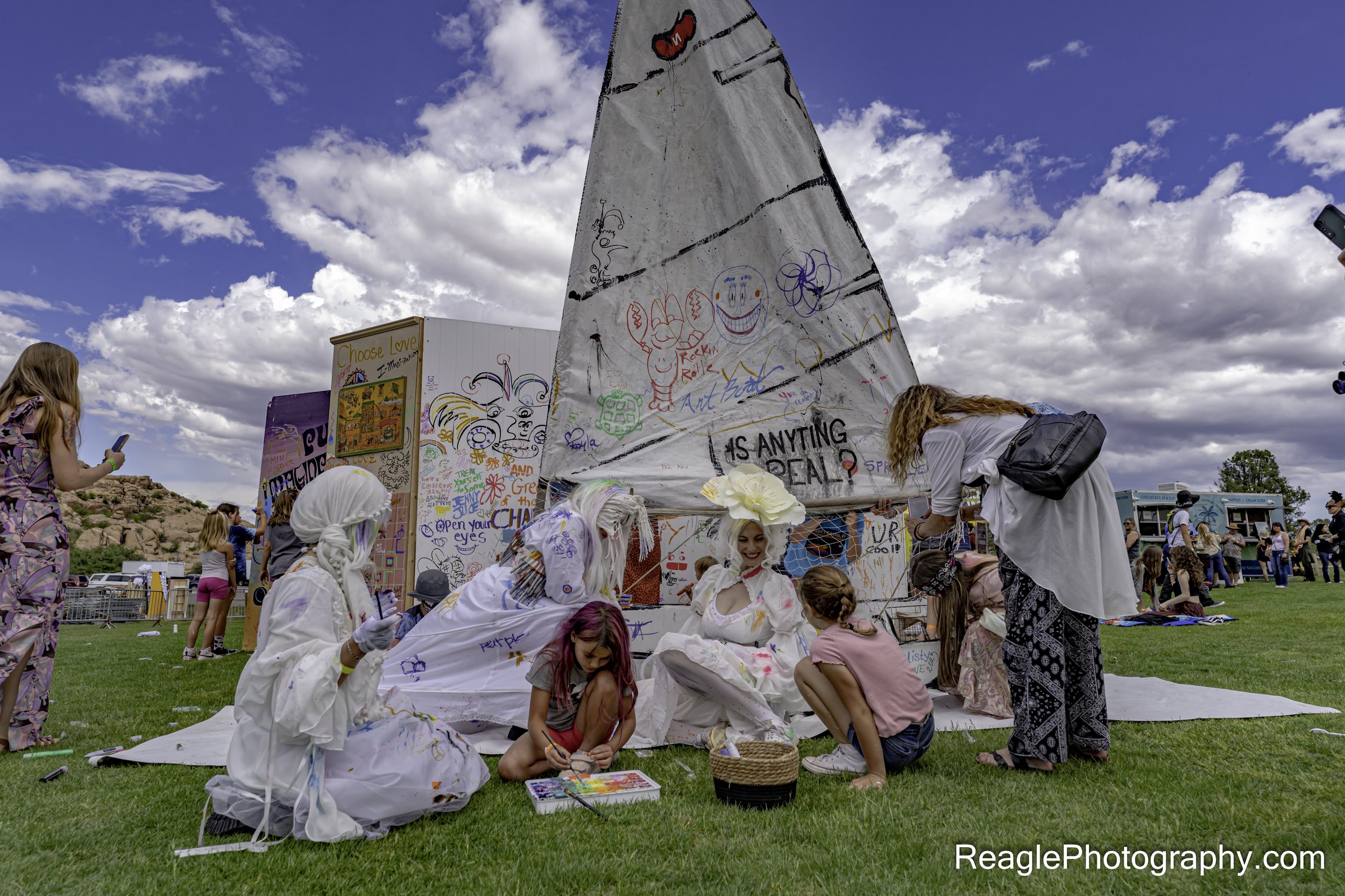 People, including children, gathered around a large white sculpture with colorful drawings and writing on it, at an outdoor event on a cloudy day.