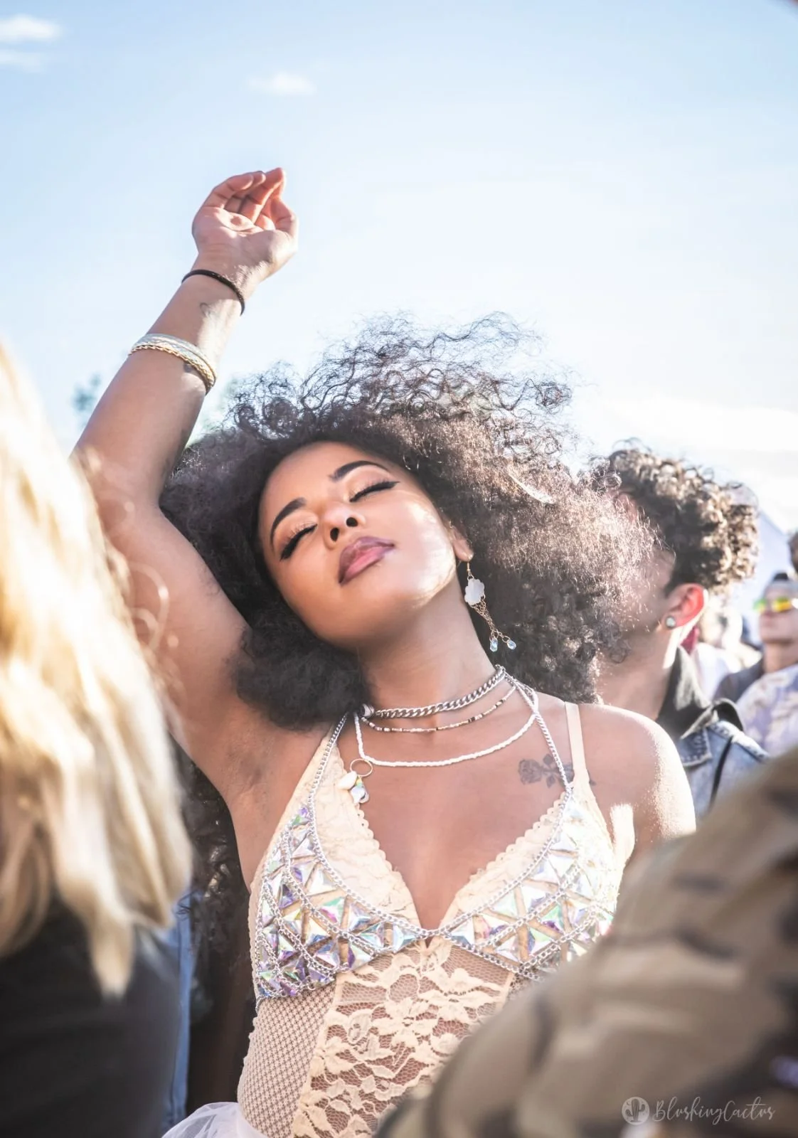 A woman with curly hair and closed eyes, raising her arm, appears to be dancing at an outdoor event under a blue sky.