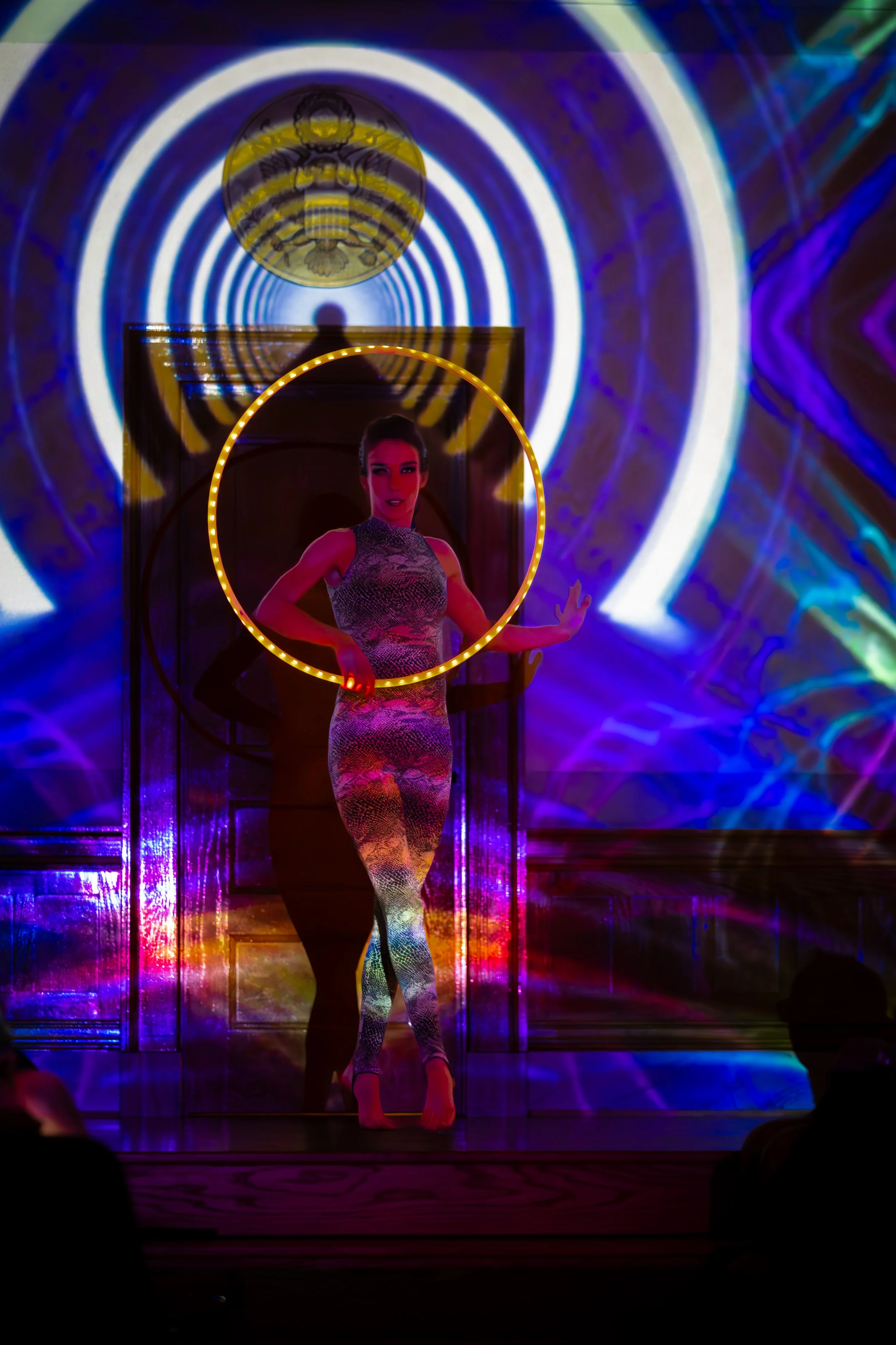 A performer standing behind a circular light ring with a colorful, abstract light display projected on the background and floor.