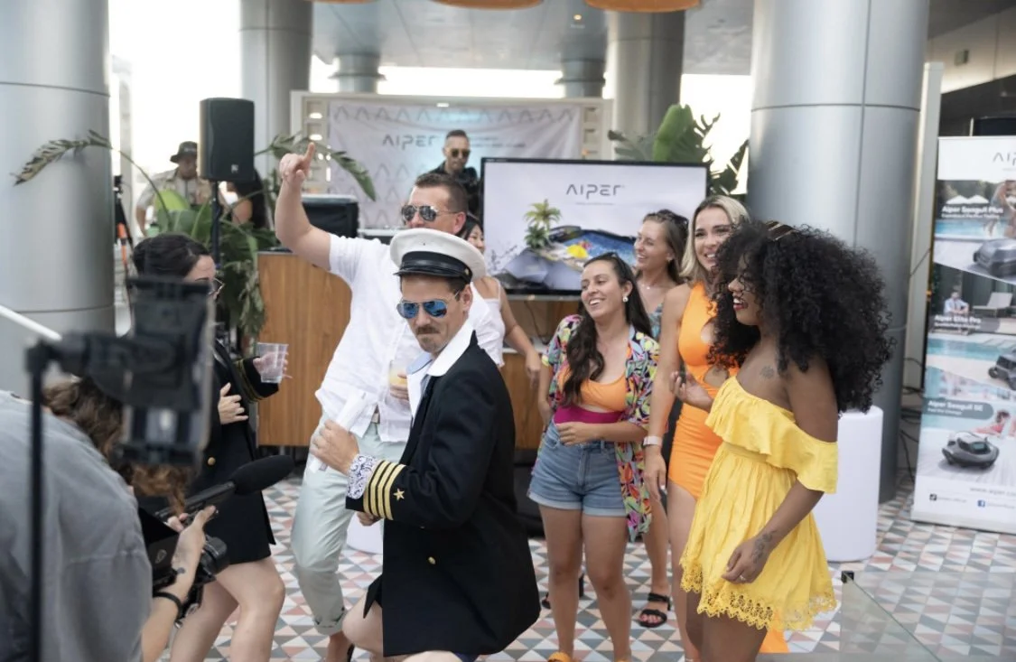 Group of people at a party, some dancing and smiling, with a DJ in the background and promotional banners nearby.