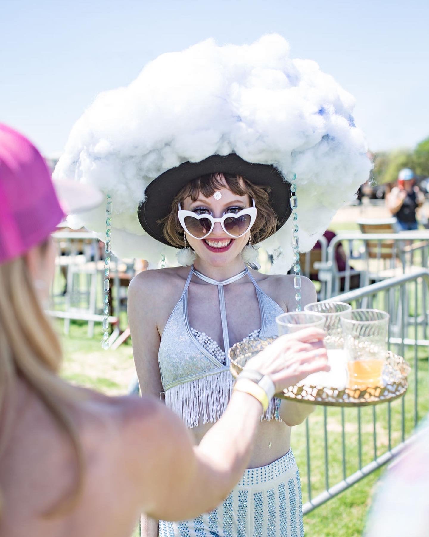 Woman wearing a large cloud-shaped hat, heart-shaped sunglasses, and a sparkly silver top, smiling and holding drinks at an outdoor event.