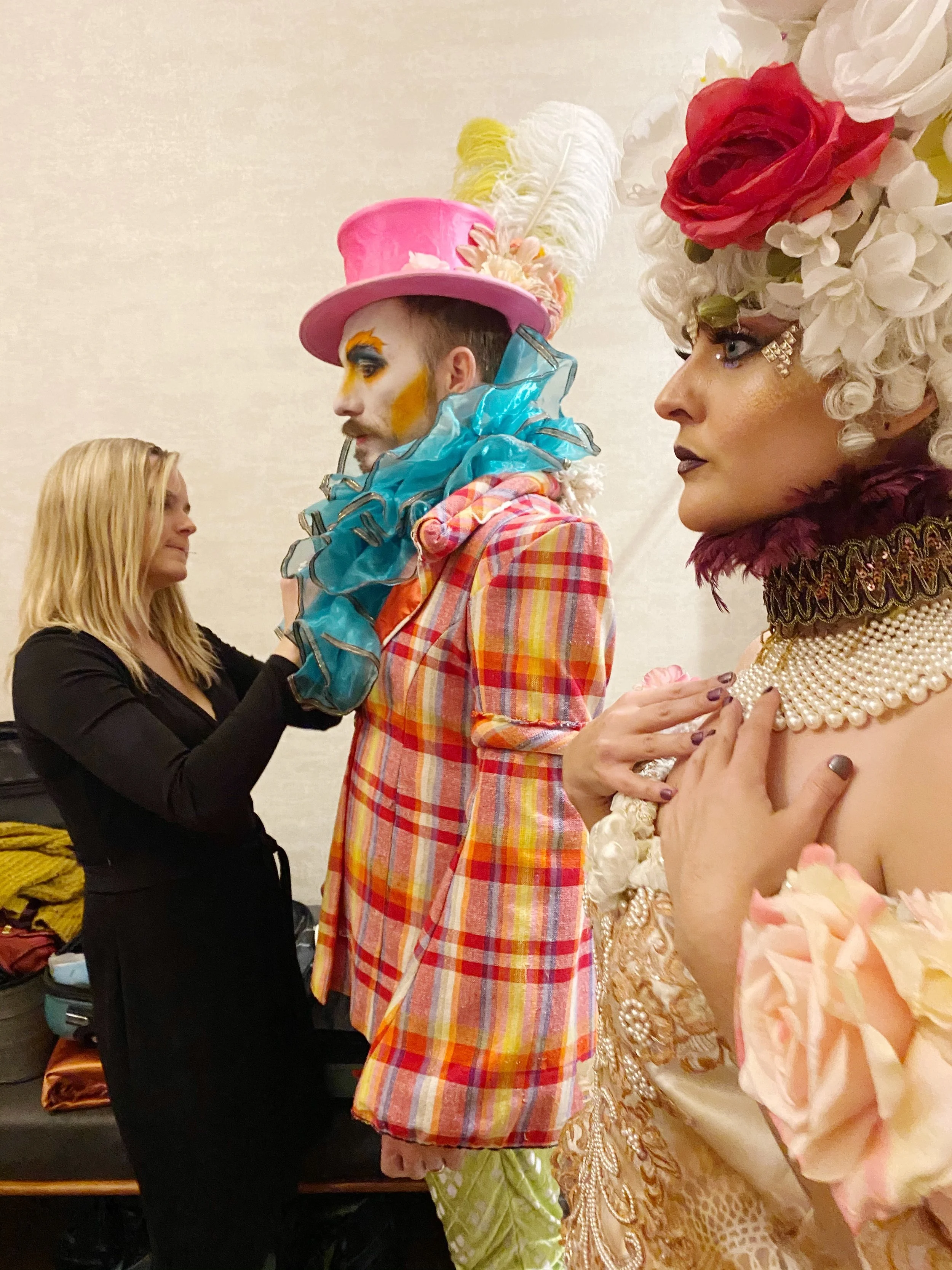 A performer in colorful, flamboyant costume and makeup is being helped by a woman backstage, with another performer in elaborate costume and headpiece watching.