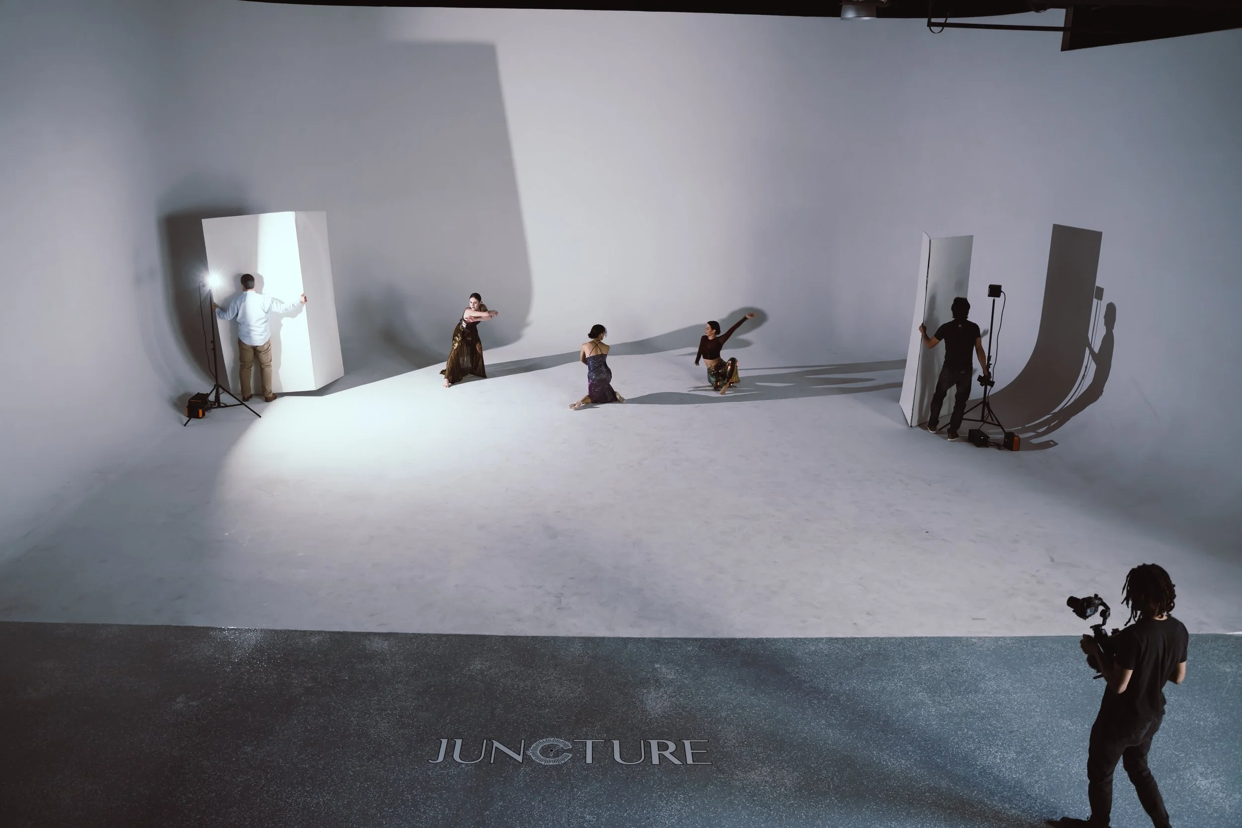 A photo of a photography studio with three models in the middle, two photographers on each side with lighting equipment, and a cameraman in the foreground. The studio has a white backdrop, and the word "JUNCTION" is visible on the floor.