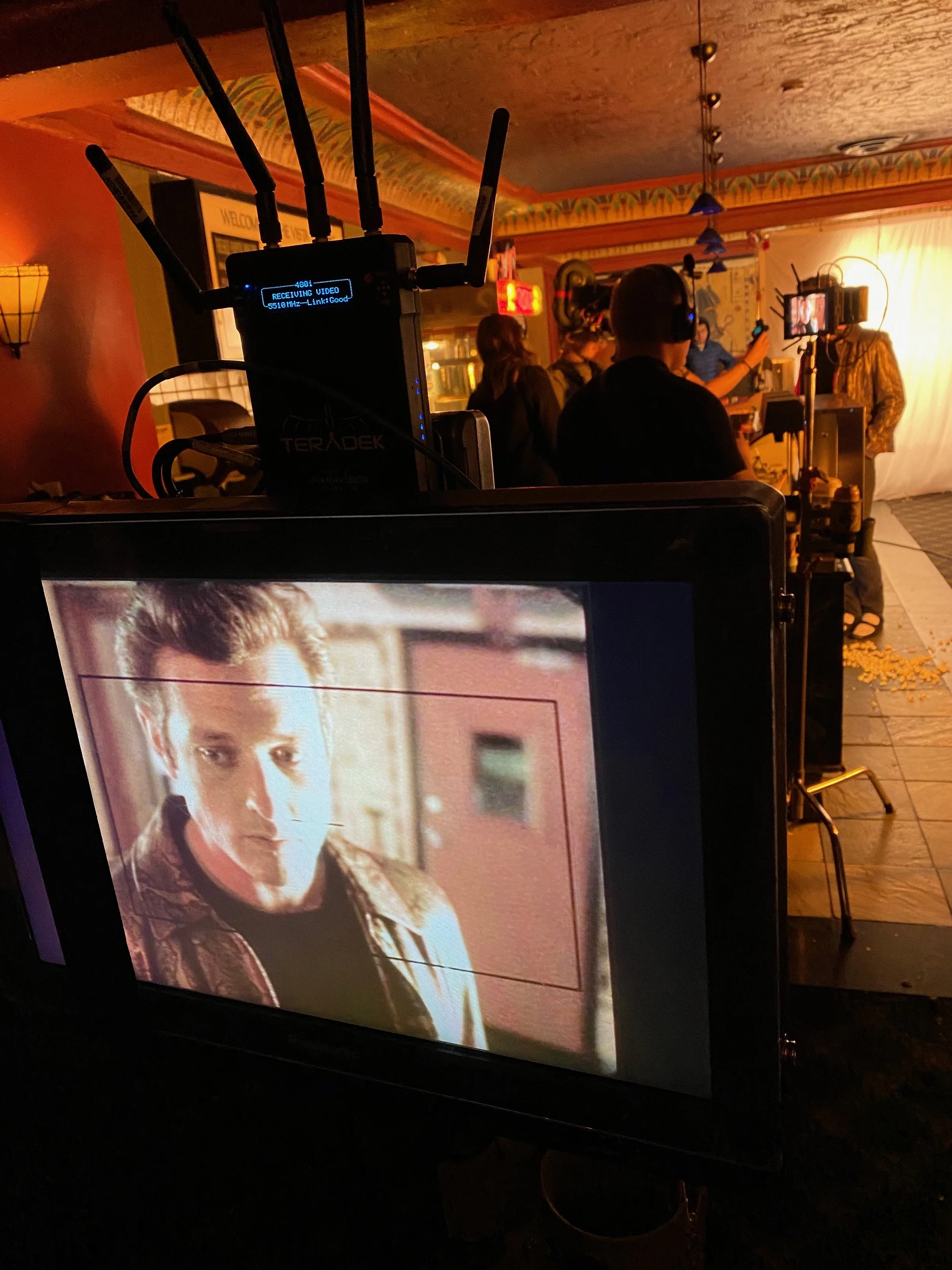 A behind-the-scenes shot of a film or video shoot. The monitor screen in the foreground shows a close-up of a person's face. In the background, people are gathered around with filming equipment, including a camera on a tripod, set up in a colorful, d