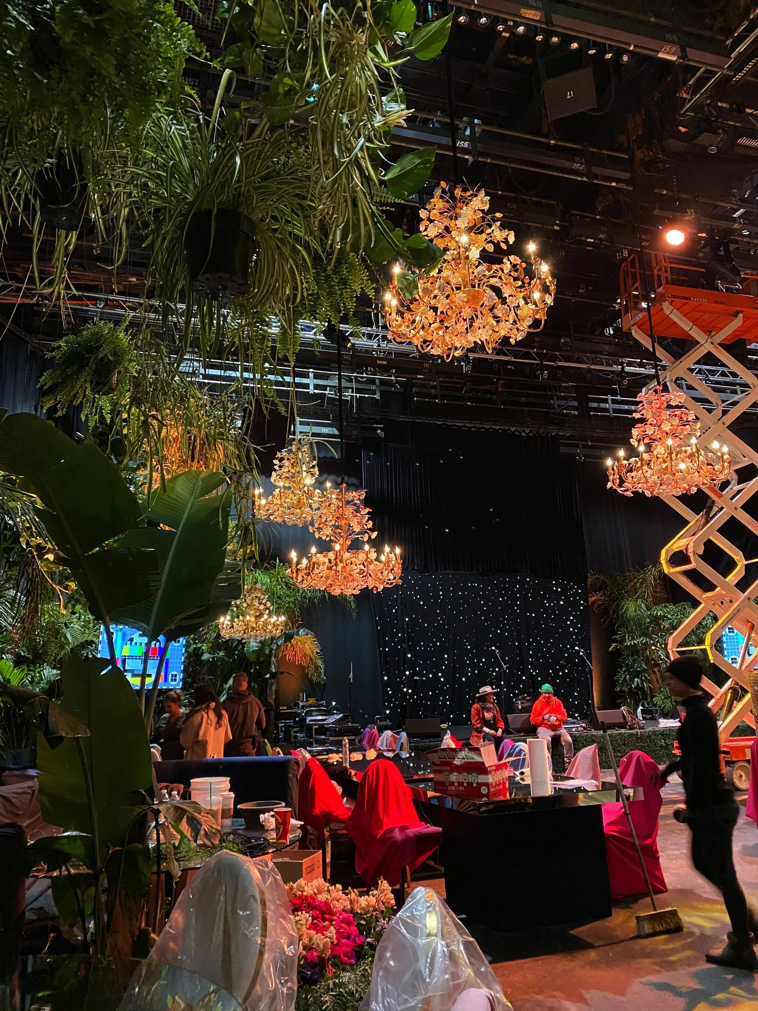 A stage decorated with hanging chandeliers and surrounded by lush green tropical plants. There are people preparing and setting up for an event, with chairs covered in red and pink cloths and tables with various items. The scene appears to be inside 