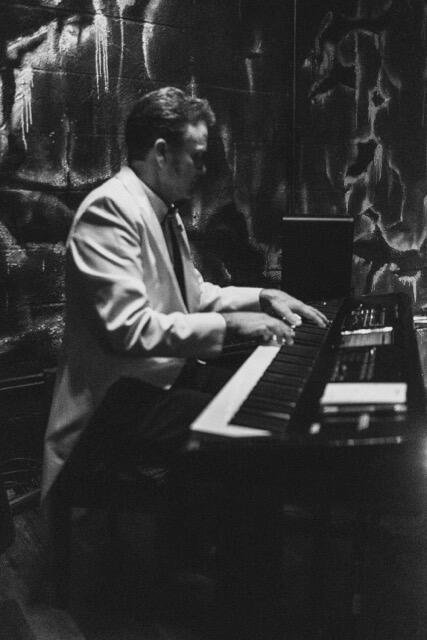 A man in a light-colored suit playing a piano in a dimly lit room with textured walls.