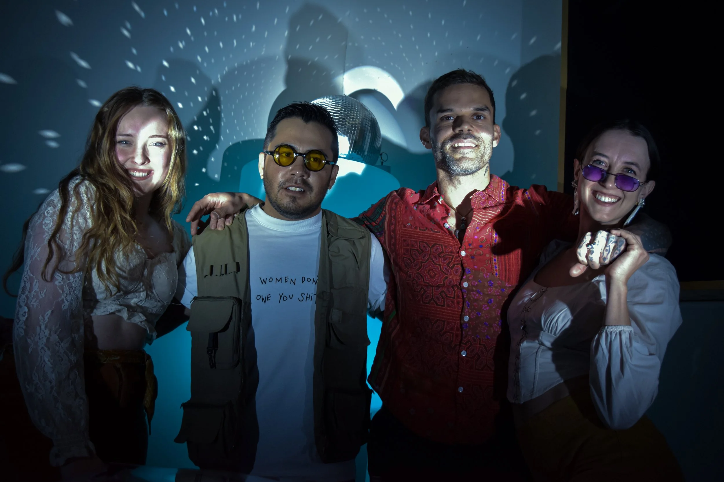 Four diverse friends smiling and posing together at a party with a disco ball and light effects on the wall behind them.