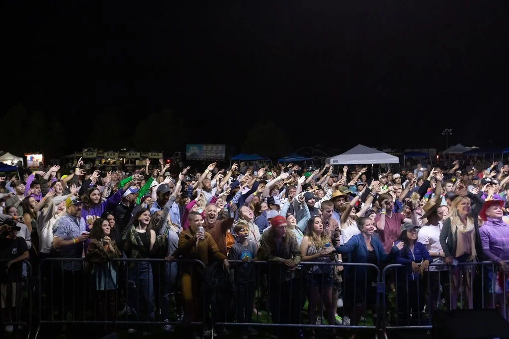 Crowd of people at a concert or festival at night, raising their hands and enjoying the performance, with tents and stalls in the background.