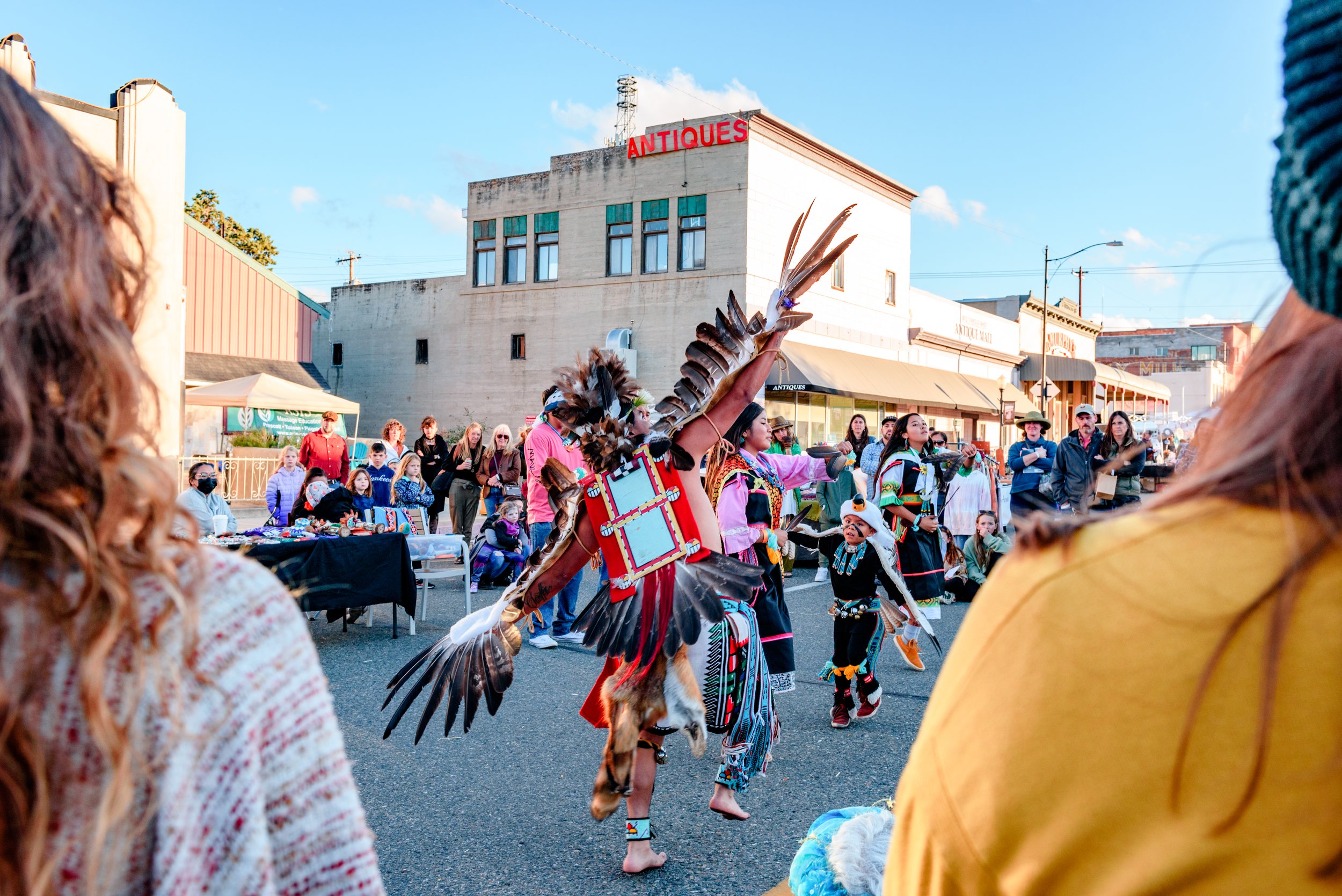 Native American dancers performing in a street parade with spectators watching under a clear blue sky.