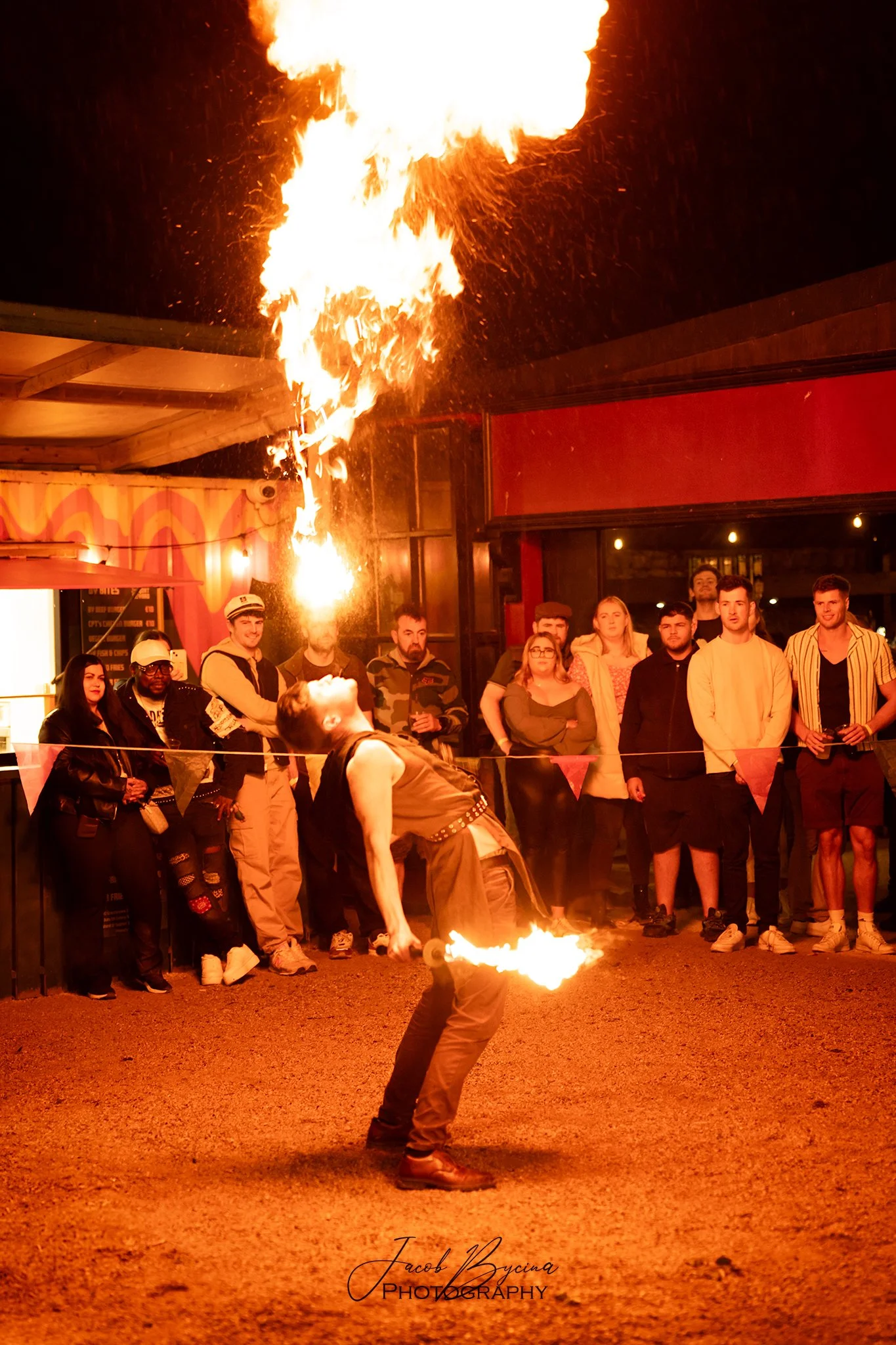 A fire performer is shooting flames while performing for an audience at night. The performer is leaning back, blowing fire from his mouth, with flames extending upwards and downwards. A crowd of spectators watches the performance behind a rope barrie