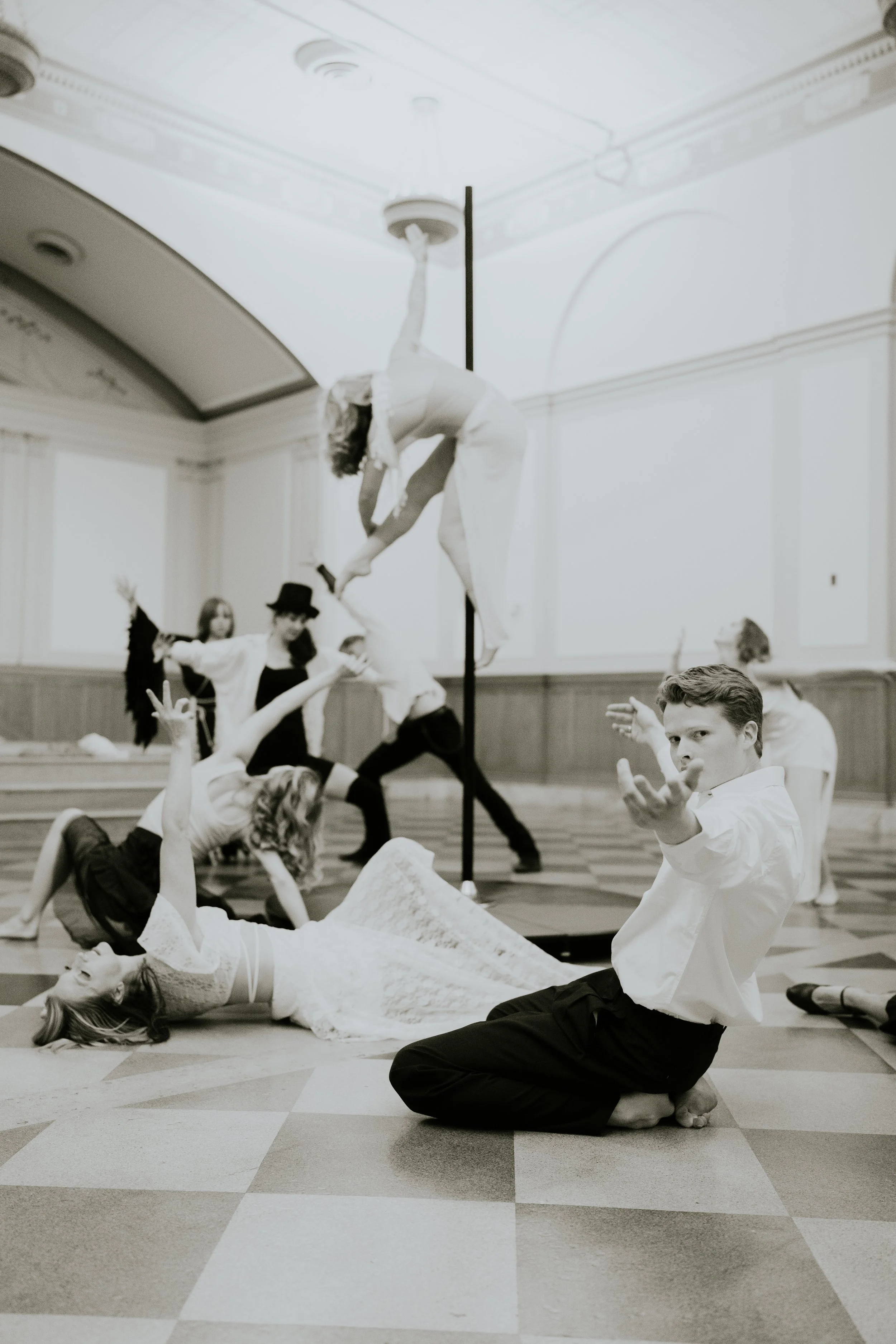 A group of people performing a theatrical or dance act on a checkered floor in a large, elegant room. One person is under a pole, hanging upside down, while others are on the ground in expressive poses. The scene appears dramatic and artistic.
