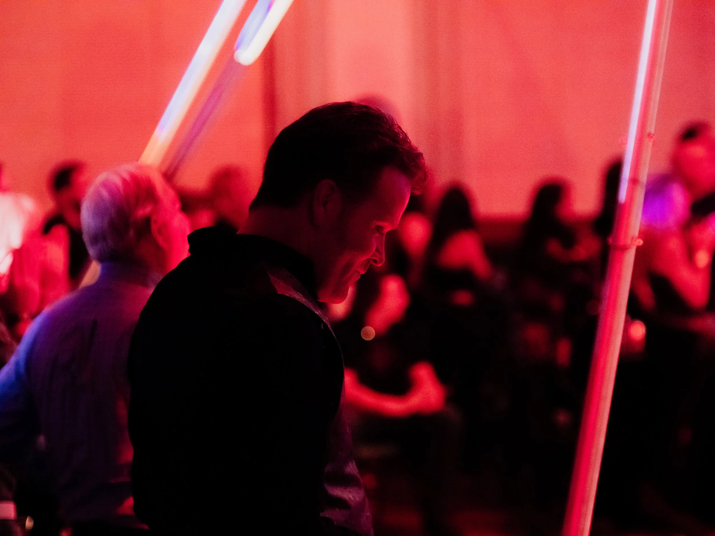 A young man smiling in a dimly lit setting illuminated by red neon lights with a crowd of people in the background.