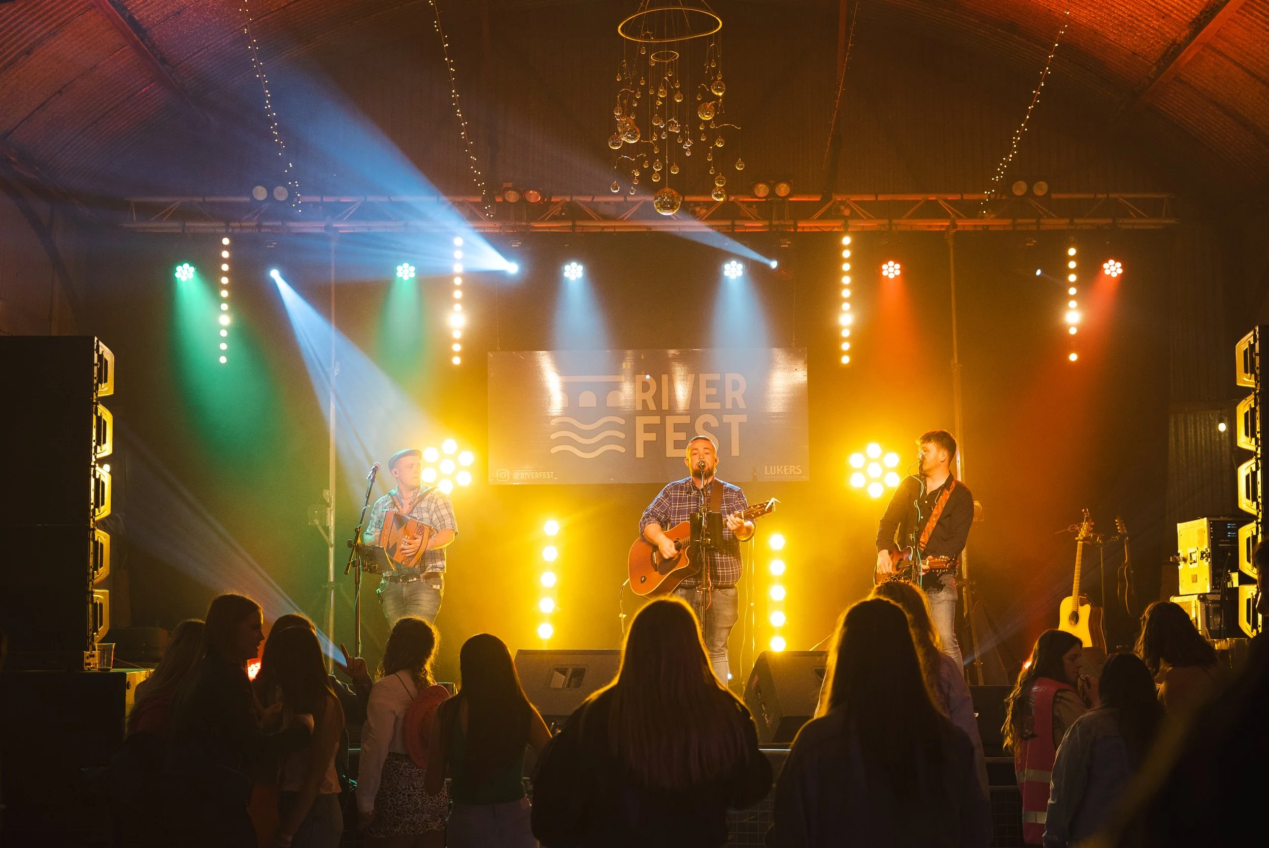 A band performs on stage at River Fest with three musicians playing guitars and singing, colorful stage lights illuminating the scene, and an audience watching in the foreground.