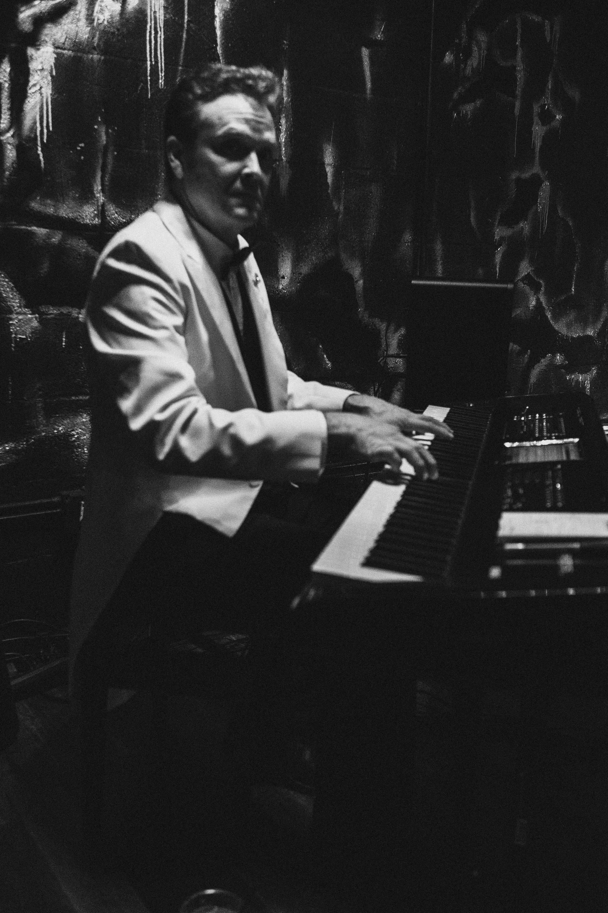 A man in a white suit playing a piano in a dimly lit setting with a textured wall in the background.