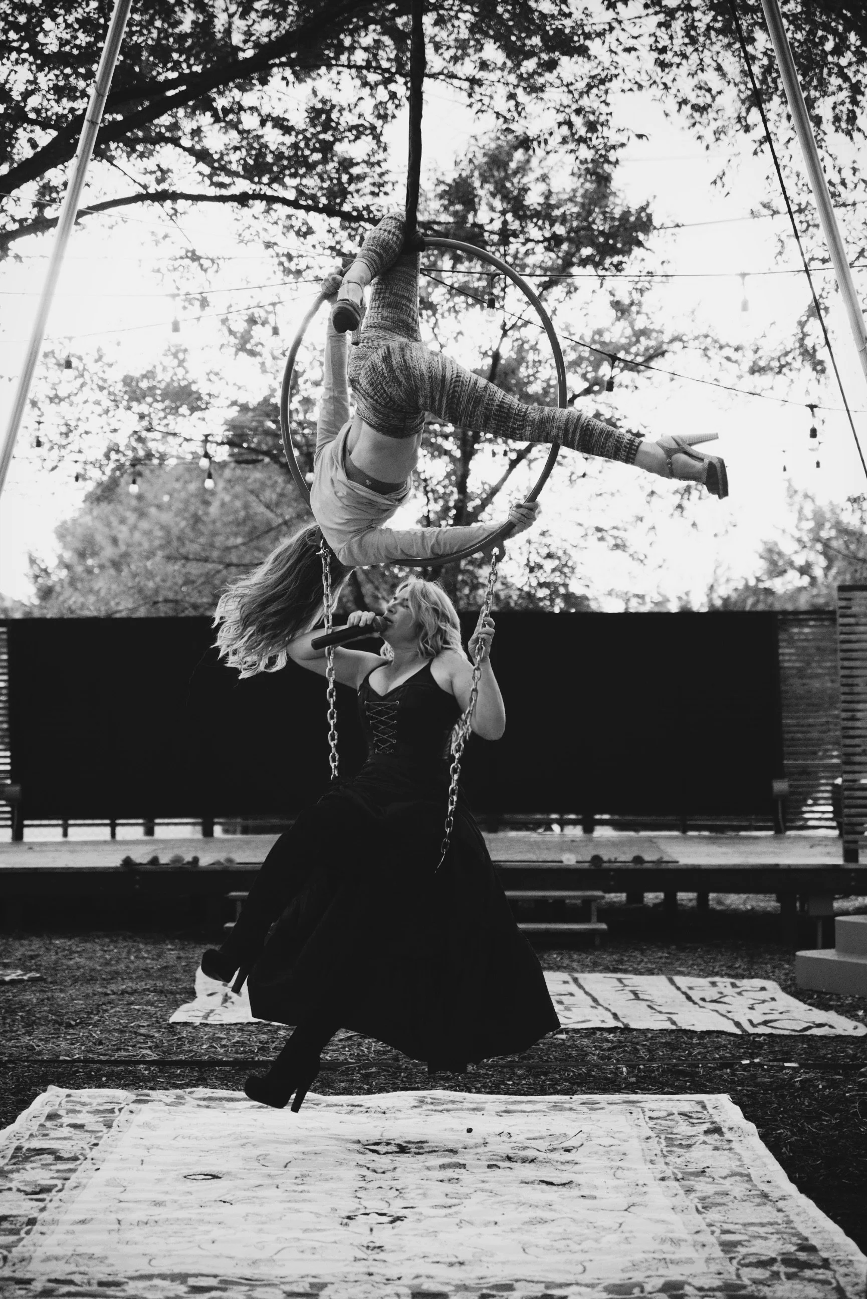 A woman singing into a microphone while sitting on a swing beneath a woman performing acrobatics on an aerial hoop outdoors with trees and string lights in the background.