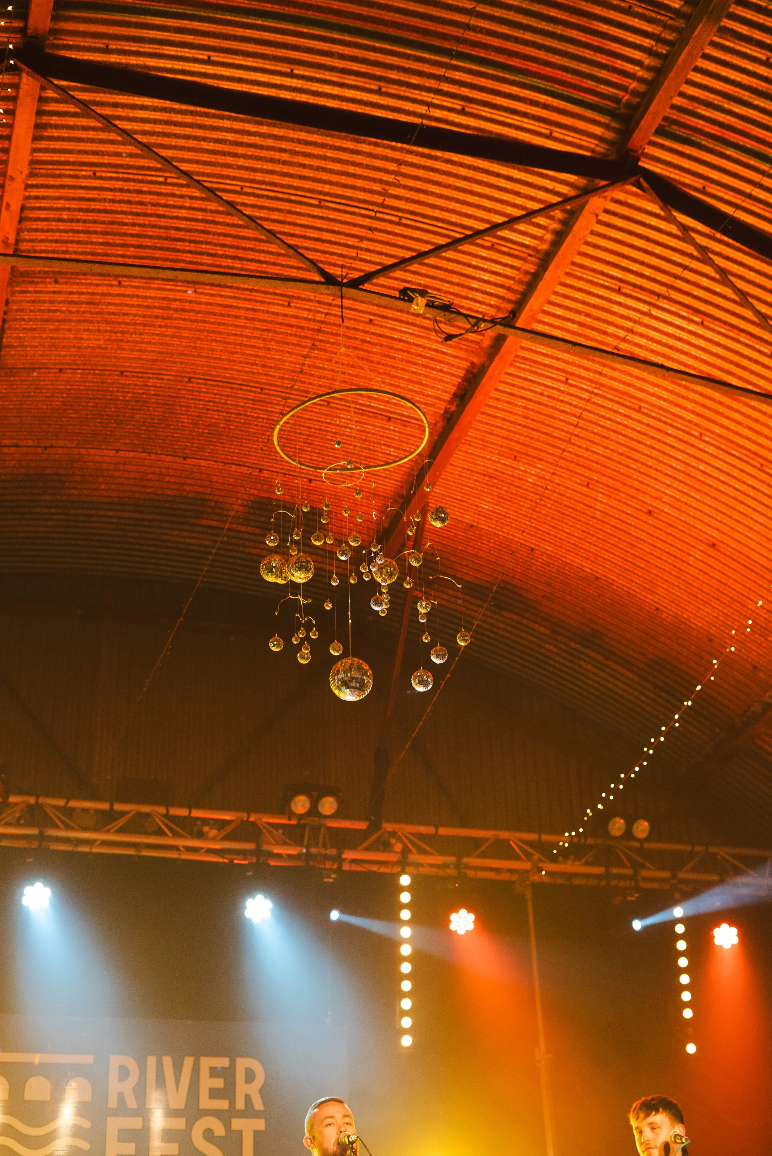 A stage at an event called River Fest with two performers singing into microphones, colorful stage lighting, and decorative hanging disco balls and ornaments.