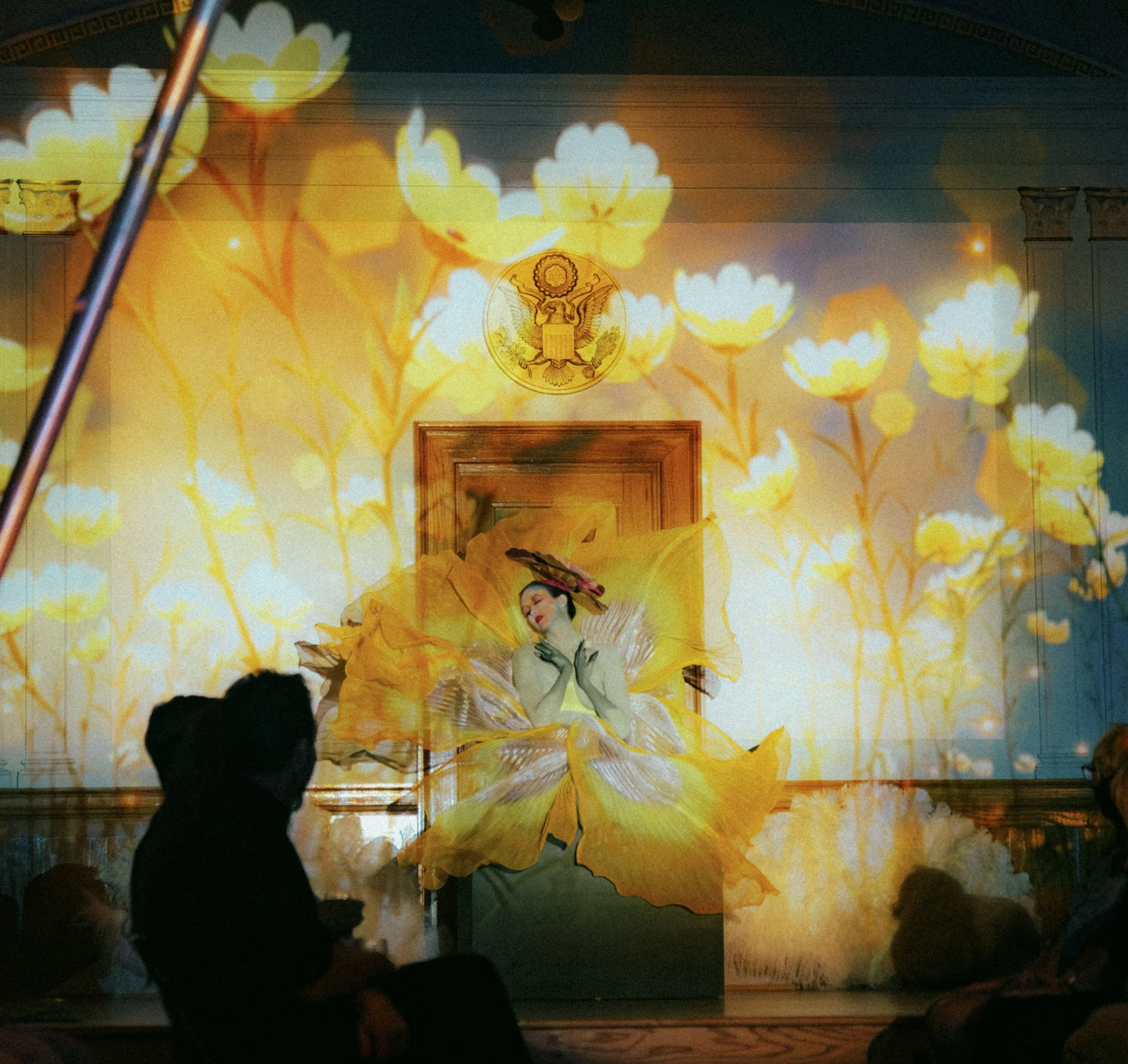 A woman performing as part of a stage show with floral projections behind her, dressed in a costume resembling a yellow flower, with a floral hat, on a stage with an ornate background and audience seated in front.