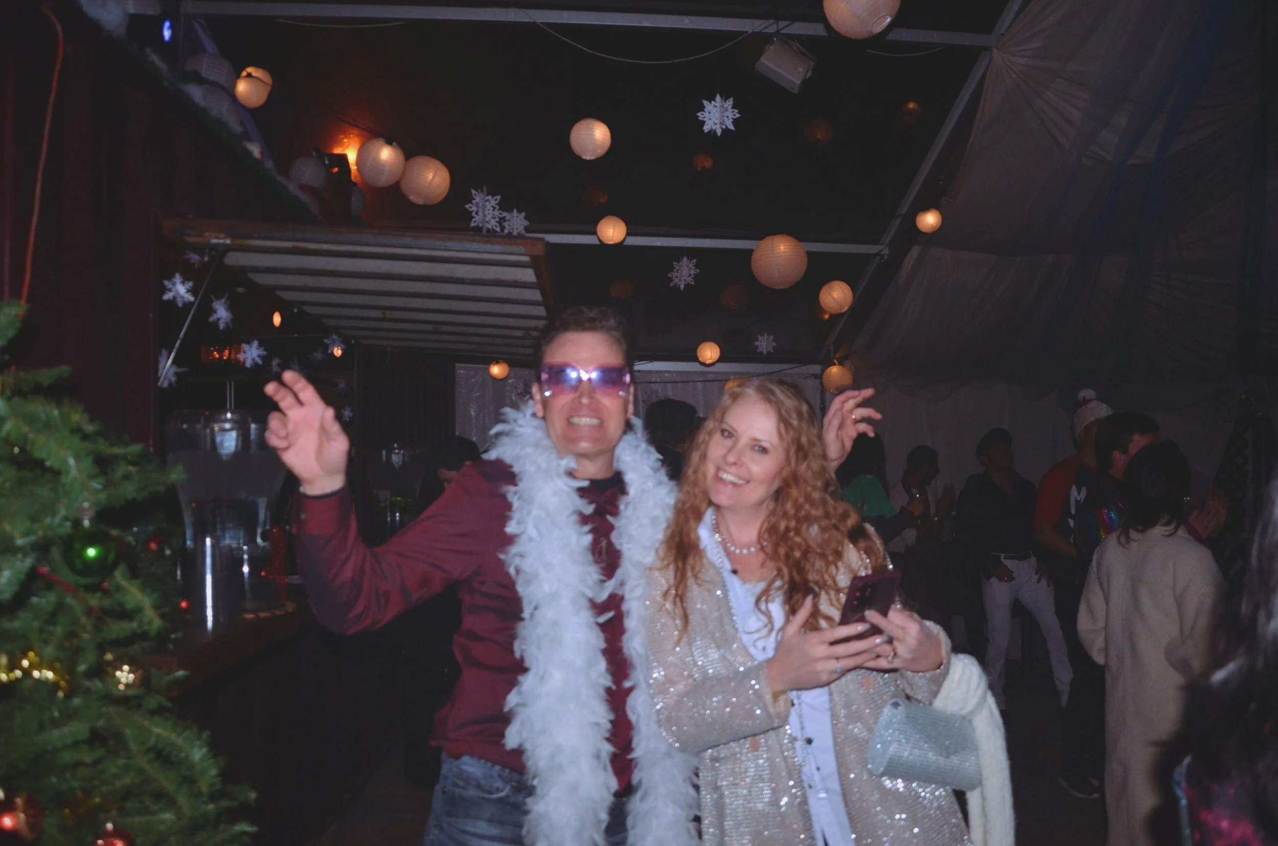 Two people dancing and smiling at a Christmas party, surrounded by holiday decorations like paper lanterns and snowflakes, with other guests in the background.