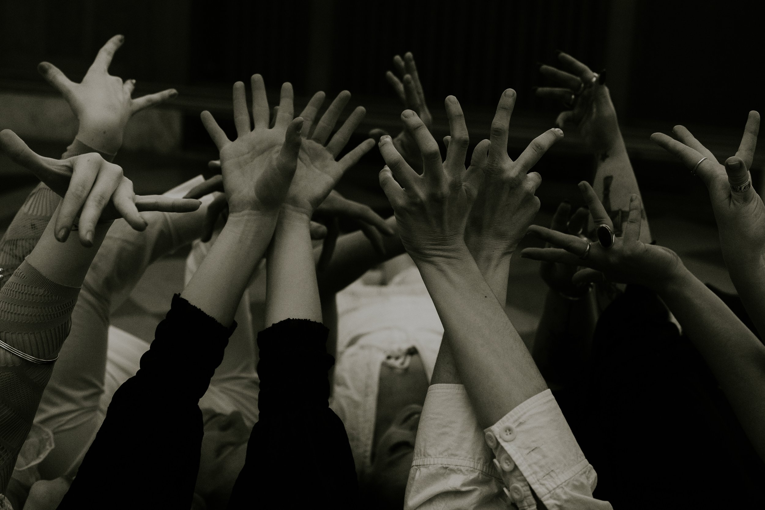 Multiple hands reaching upward, some with rings and bracelets, in a black-and-white photo.