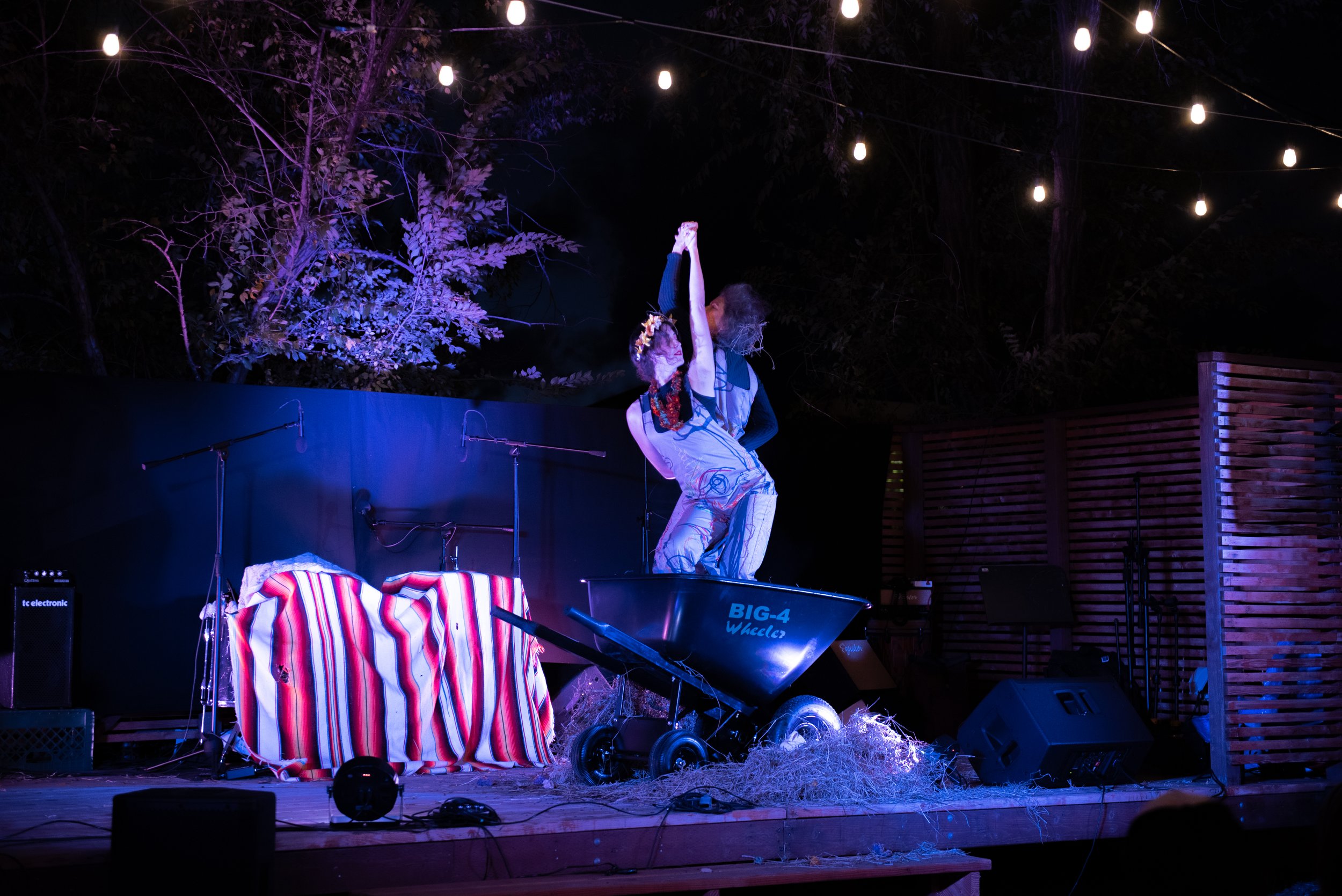 Performer dancing on stage at night with string lights overhead, surrounded by trees, with various stage equipment and decorations.