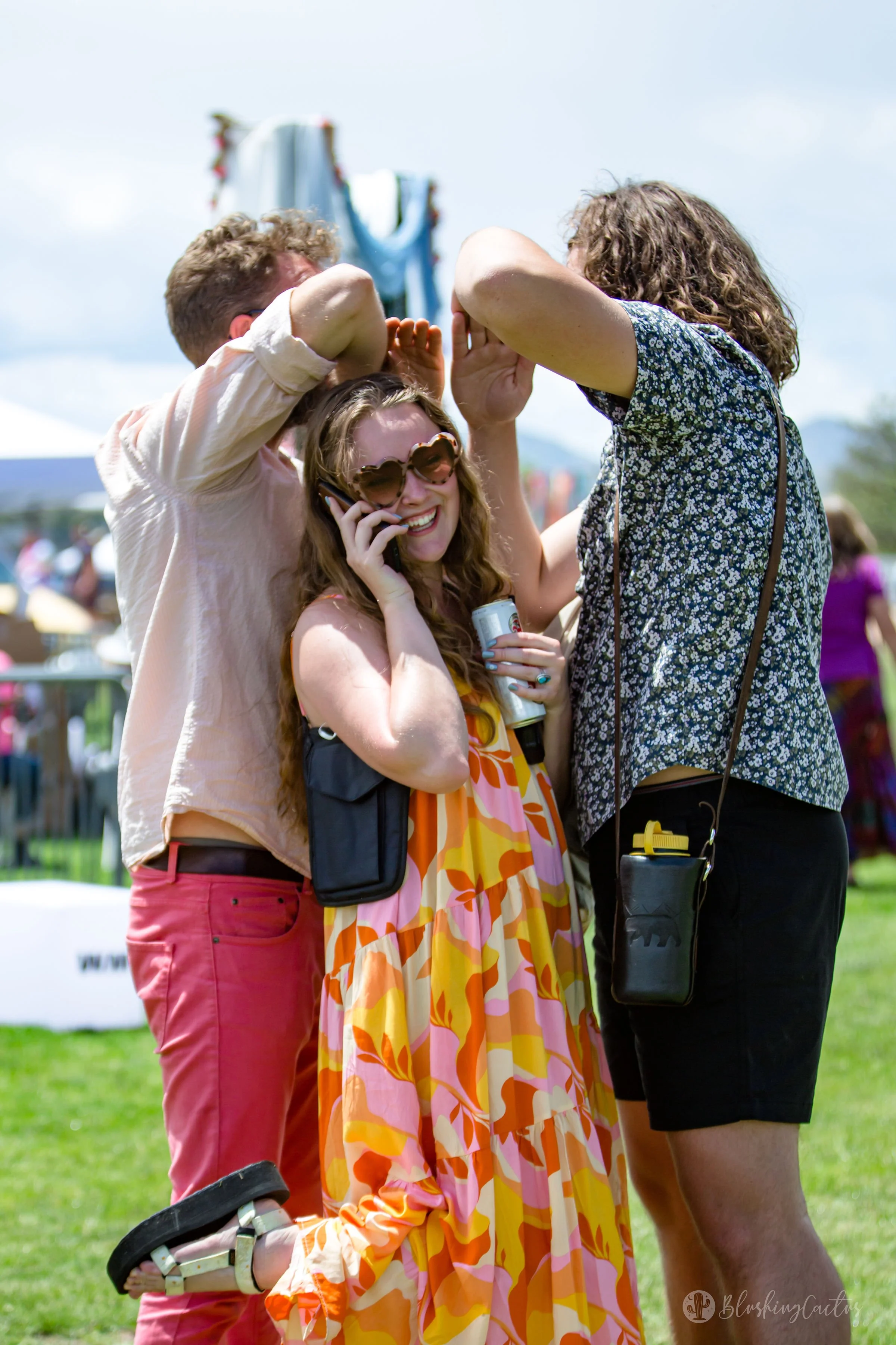 Group of people at an outdoor event, with a woman talking on her phone, smiling, and wearing sunglasses, while others are dancing or socializing in the background.