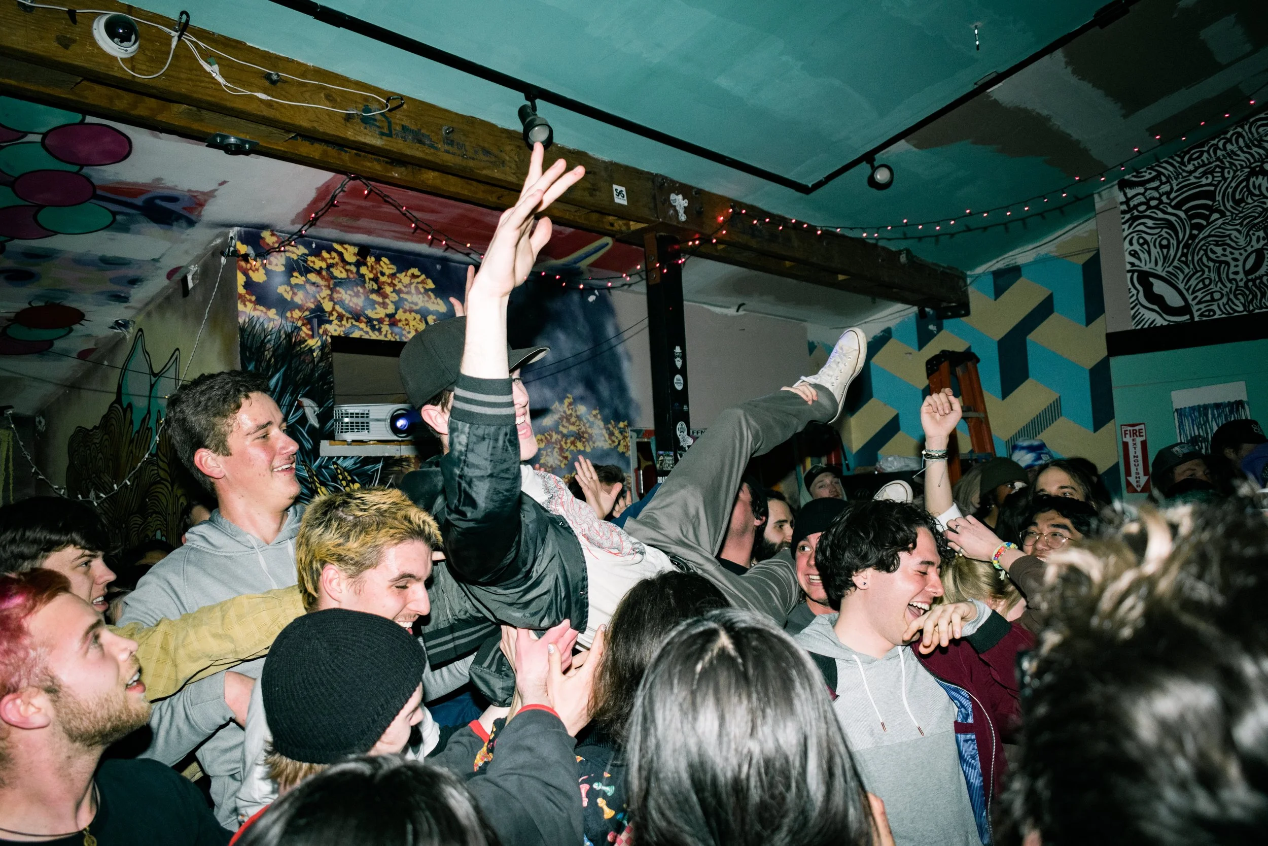 Crowd of young people at a music concert or party, with one person being lifted above the crowd and others laughing and enjoying the event.