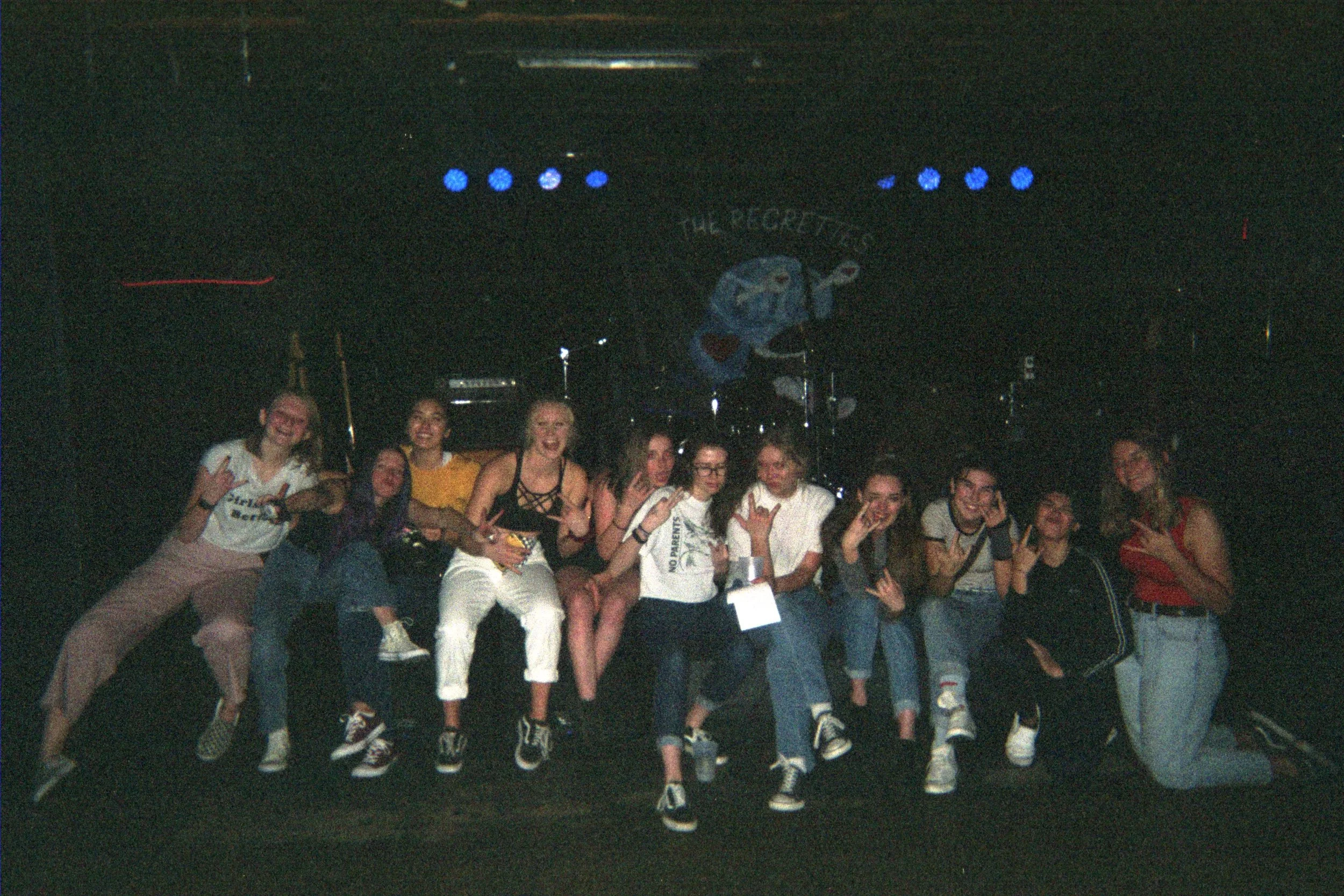 A group of people posing and making peace signs on stage with a dark background and a large mascot in the background.